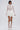 A woman poses against a plain background wearing the LUX WAVES MINI SKIRT, a sheer white lace long-sleeve top, knee-high white lace-up boots, and holding a small white handbag.