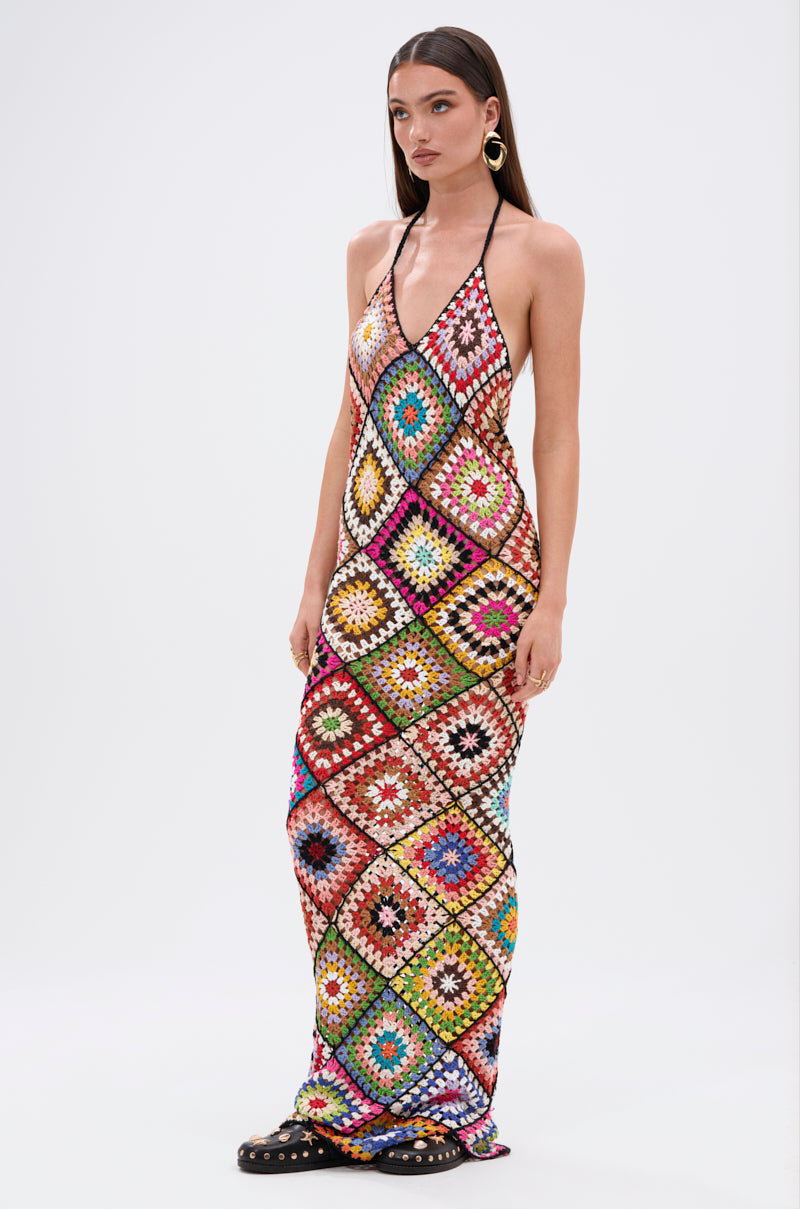 A woman wears the DONT LET ME GO CROCHET MAXI DRESS, a maxi dress with a multicolored diamond pattern, halter neckline, and open back. She completes her beach look with large hoop earrings and black sandals accented with gold studs.