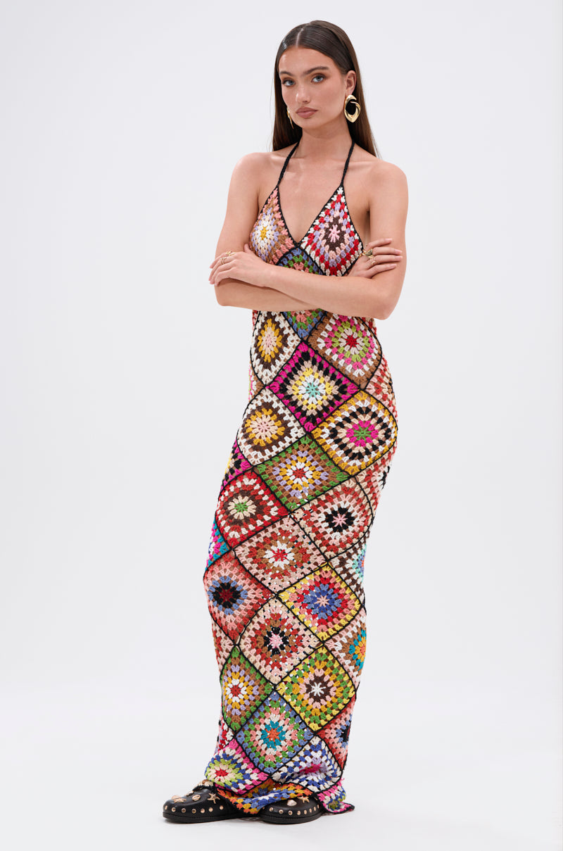 A woman stands with arms crossed in the DONT LET ME GO CROCHET MAXI DRESS, featuring a vibrant multicolored diamond pattern. She pairs it with large hoop earrings and black studded flats against a plain white background, highlighting her beach-ready look.