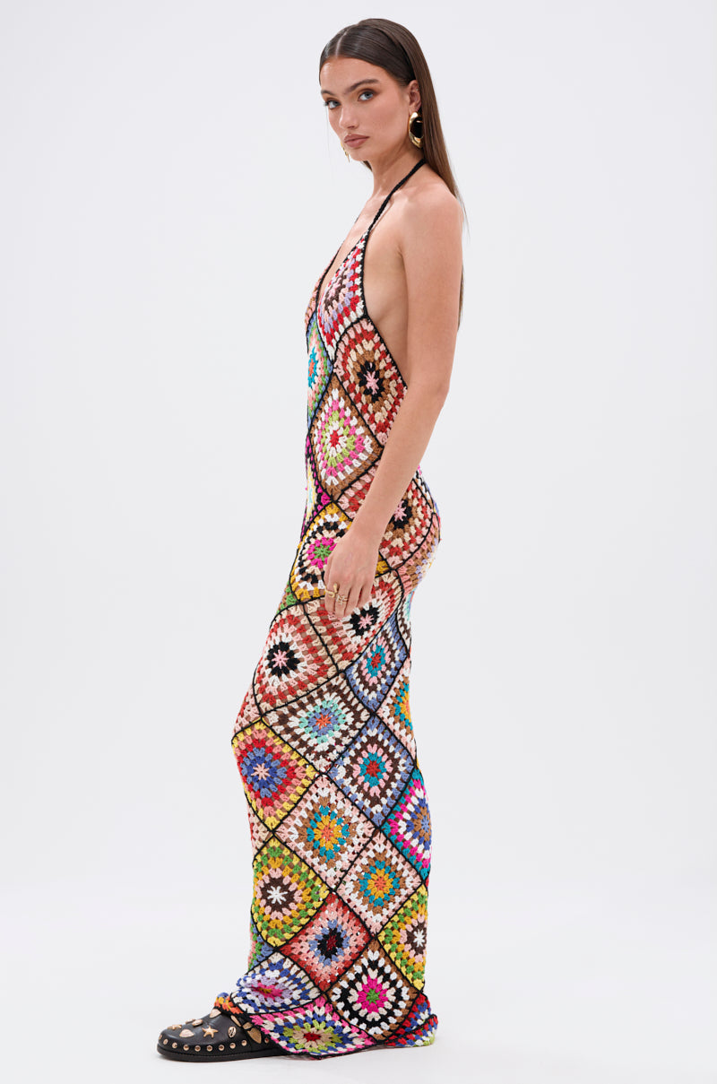 A woman with straight brown hair stands sideways against a white background wearing the DONT LET ME GO CROCHET MAXI DRESS, featuring a vibrant diamond pattern. She completes her beach-ready look with black studded sandals.