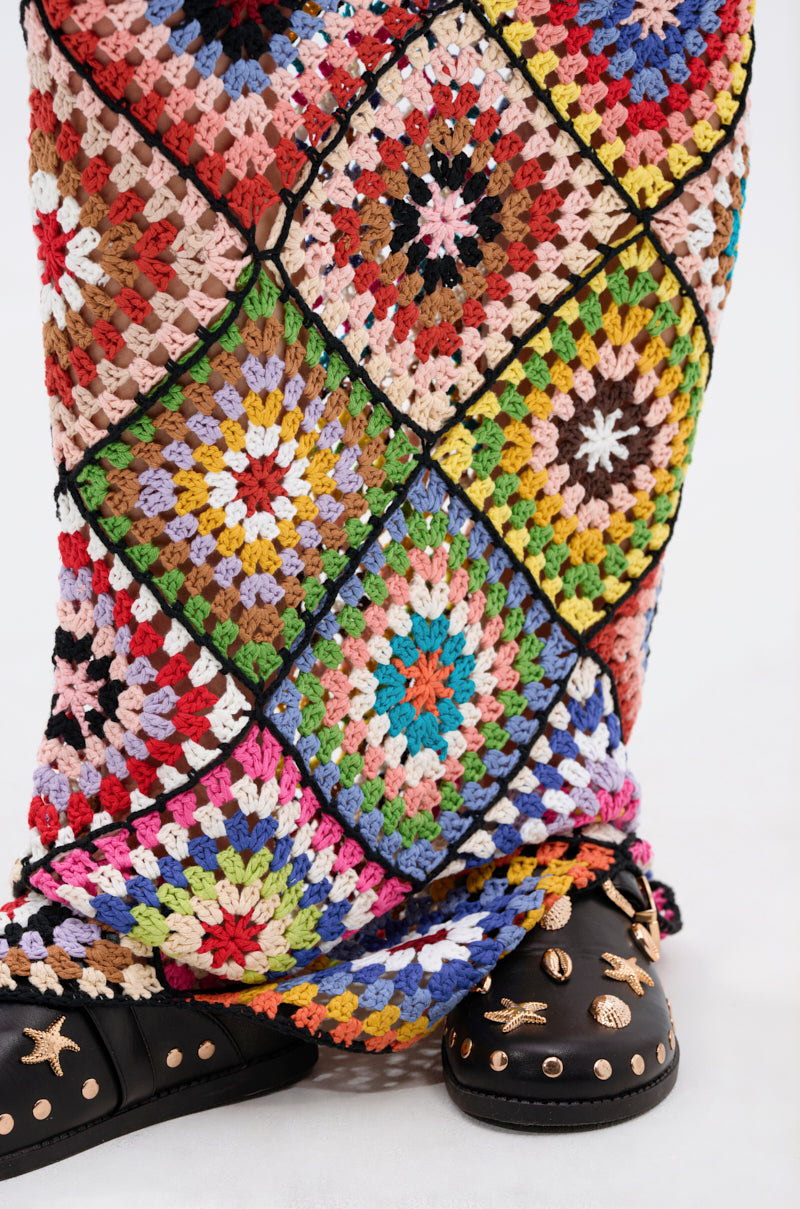 A vibrant multicolored pattern stands out in the DONT LET ME GO CROCHET MAXI DRESS featuring square designs, draped down and partly covering black shoes with gold stars, moons, and studs against a plain white background.