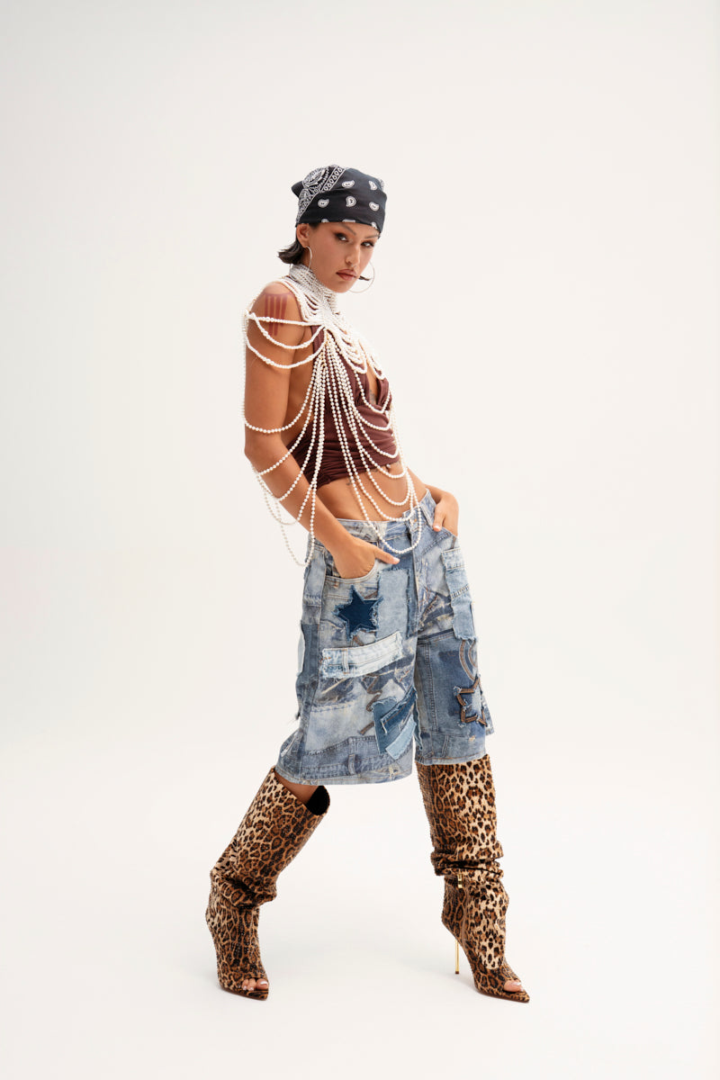 A person in a black bandana, layered pearl necklaces, and a sleeveless top exudes confidence against a plain backdrop. They're striking a pose with hands in the pockets of their "STARS IN THE SKY DENIM SHORT" paired with knee-high leopard print boots.