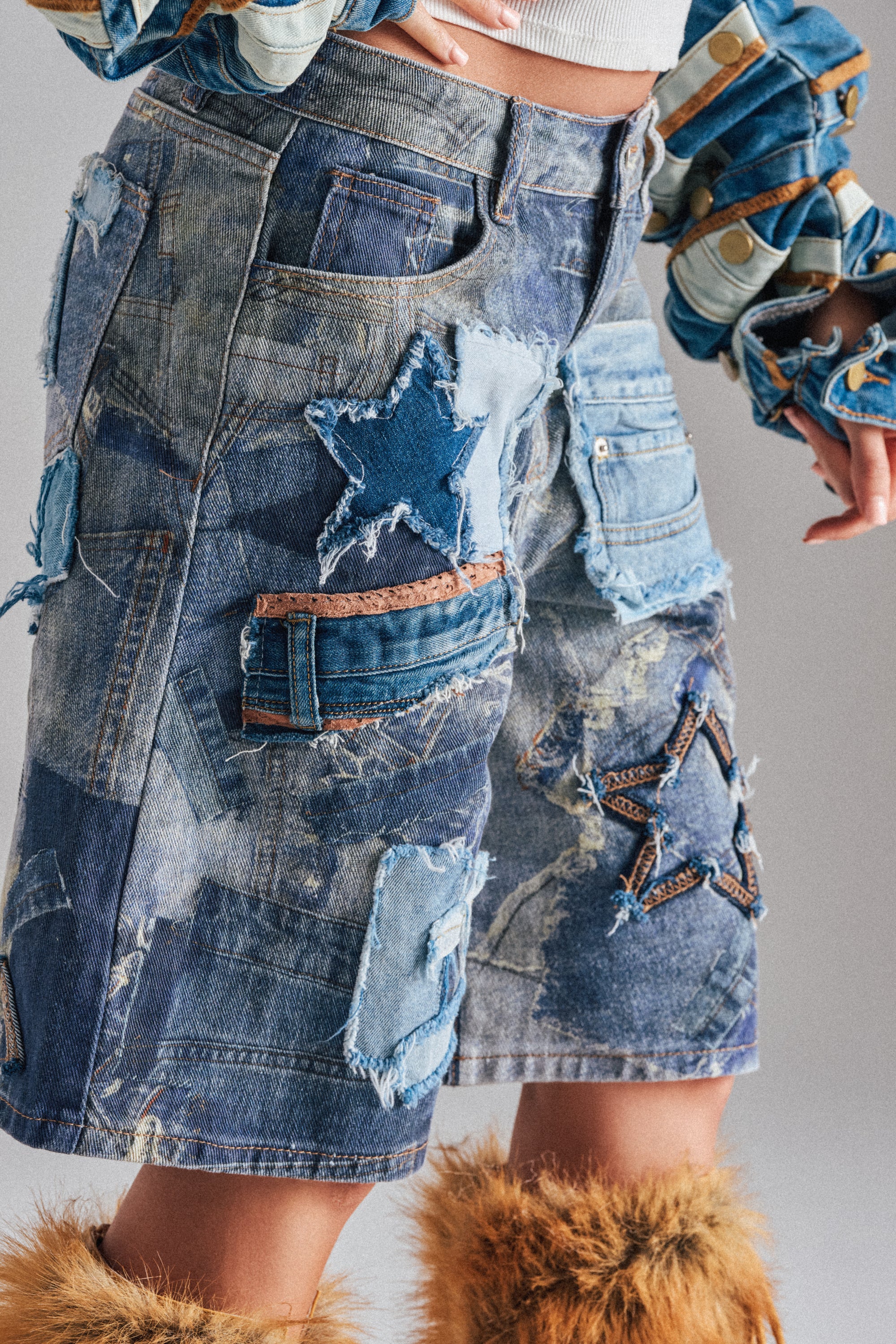 Wearing the STARS IN THE SKY DENIM SHORT with embroidered stars, patchwork denim details, layered distressing, and denim pockets, paired with furry boots and a blue jacket with round accents; only the lower body is shown.