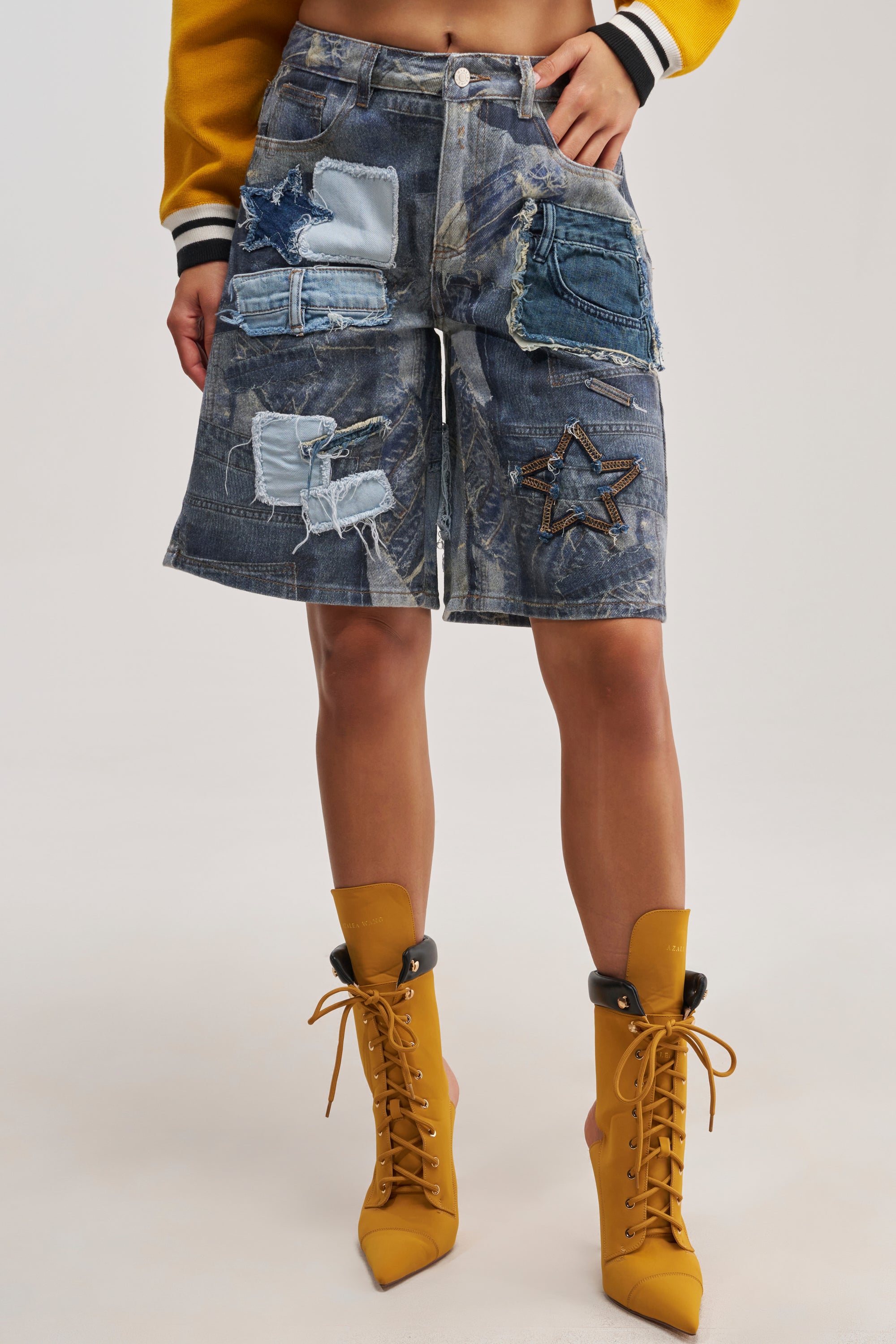Wearing the STARS IN THE SKY DENIM SHORT, featuring a patchwork of denim in various washes with star motifs, styled with a yellow and black jacket and mustard yellow lace-up heeled boots—this bold streetwear ensemble is perfect for festival season. Only the lower half of the body is visible.