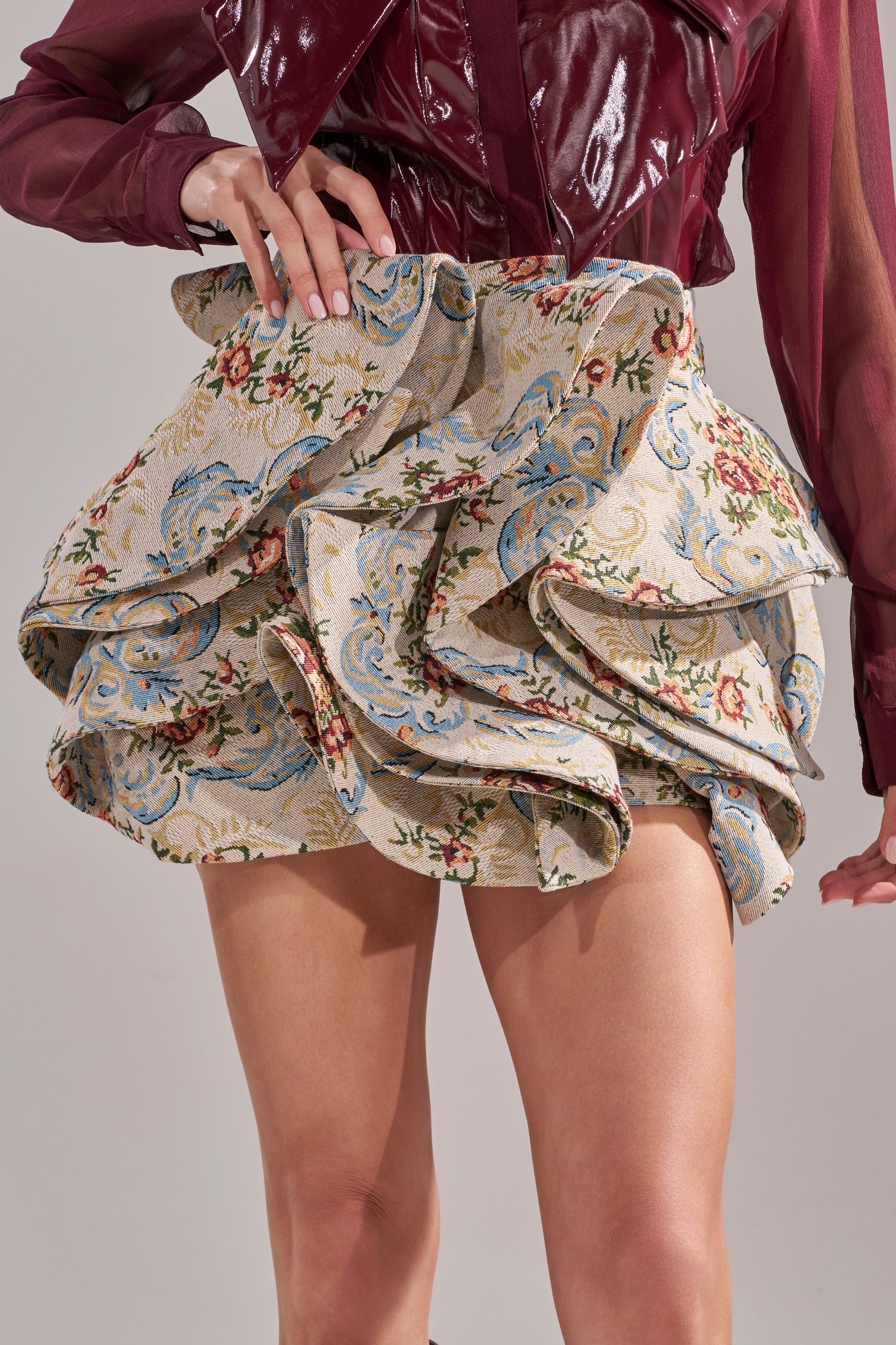 A person poses confidently with one hand on their waist, wearing a burgundy, glossy top and the FLORAL BABY RUFFLE SKIRT, which features short ruffles and blue, beige, and red floral tapestry patterns. 0