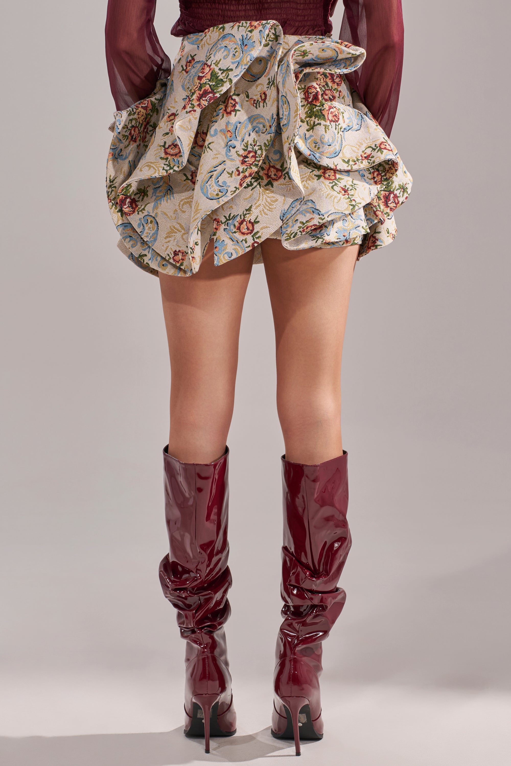 A person facing away from the camera wears the FLORAL BABY RUFFLE SKIRT with a large bow, paired with shiny knee-high burgundy boots, set against a neutral background. 4