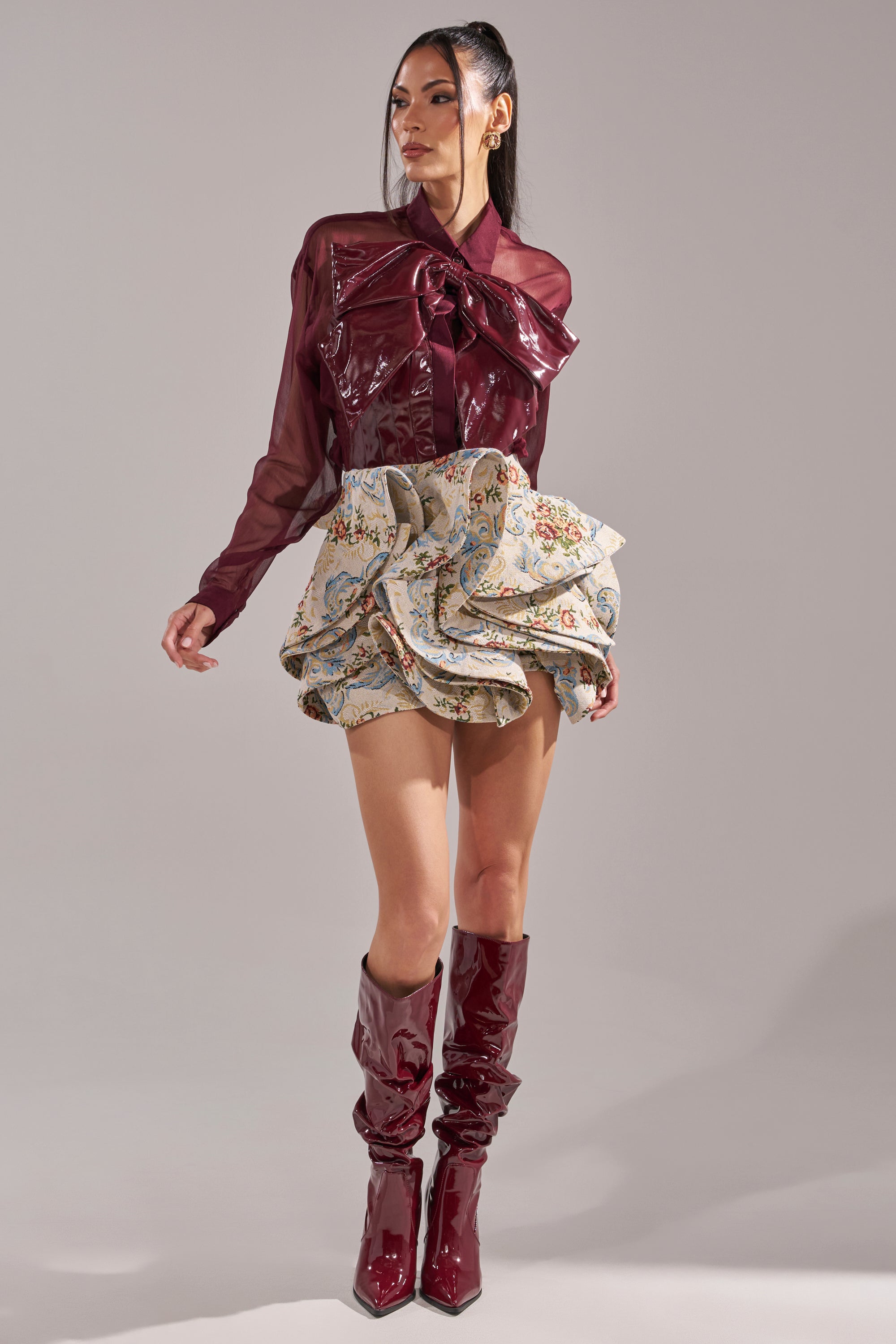 A woman models the FLORAL BABY RUFFLE SKIRT with a shiny burgundy long-sleeve blouse, large bow, and matching knee-high boots against a plain gray backdrop. 7