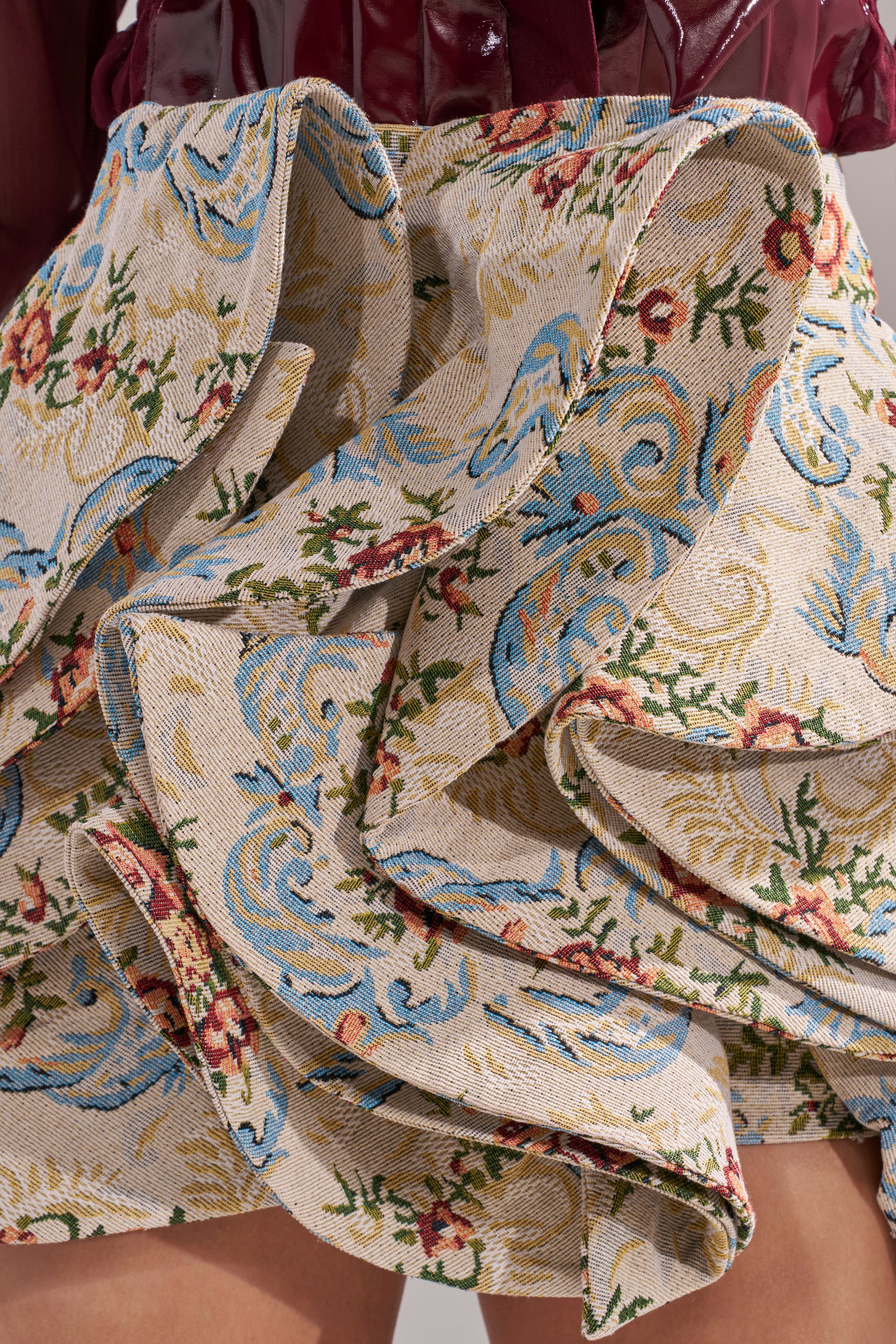 The FLORAL BABY RUFFLE SKIRT is a jacquard mini skirt featuring floral and swirl tapestry patterns in beige, blue, green, and red with ruffled details, styled here with a glossy dark red top. 8
