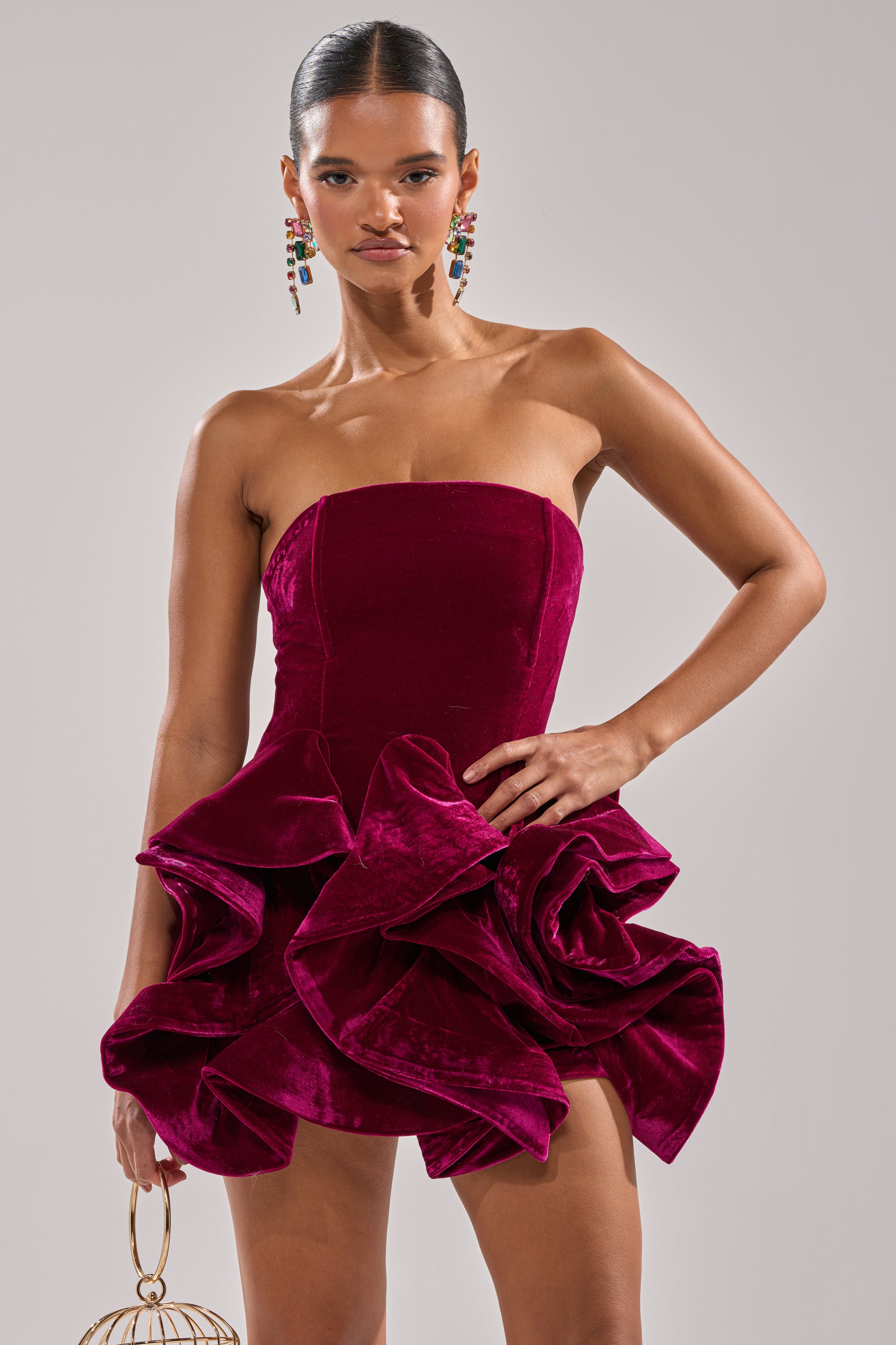 A woman wears the LOVE IS REAL VELVET MINI DRESS, strapless with dramatic ruffled skirt, accessorized with a gold sphere clutch and bold earrings, standing before a plain light backdrop.