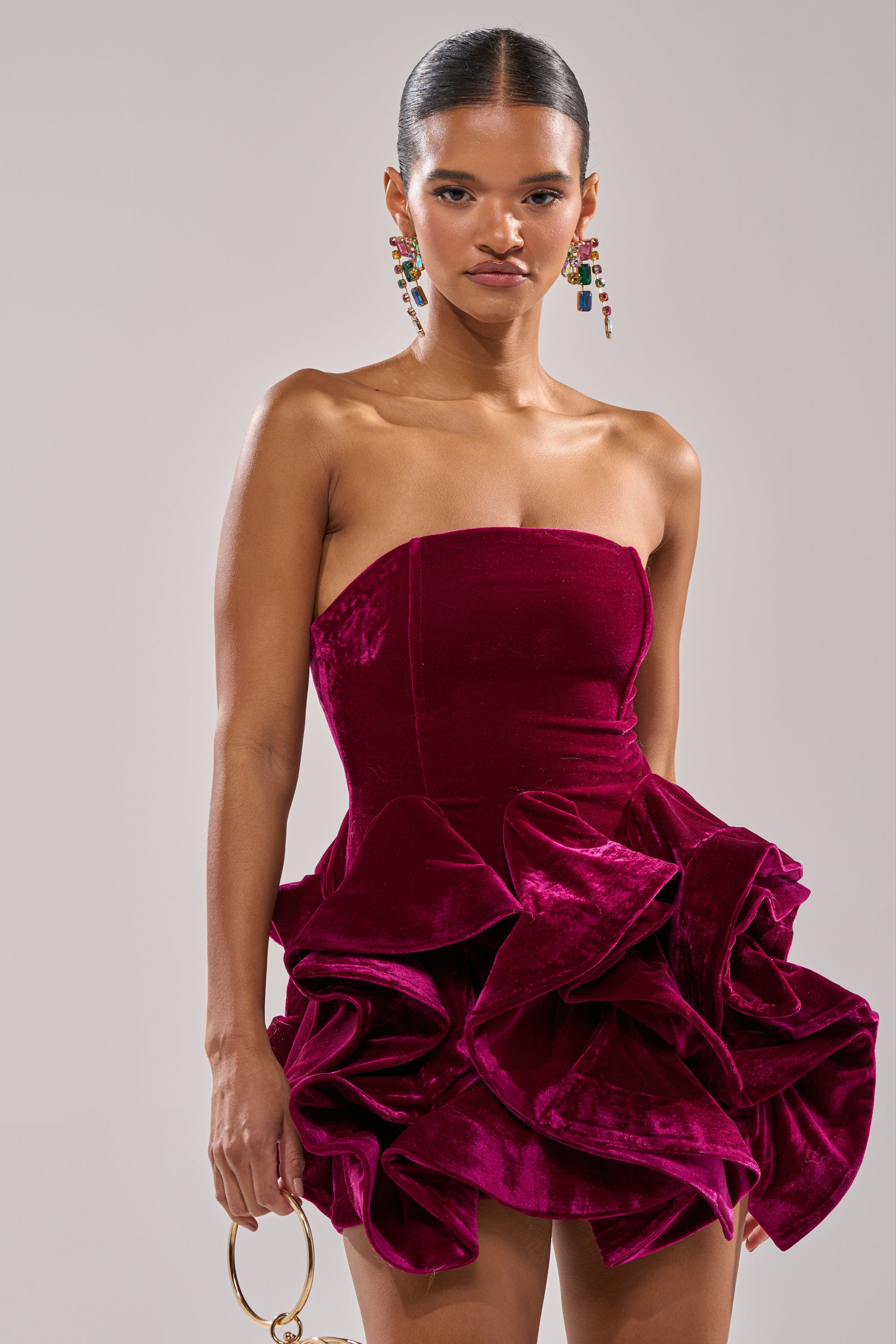 Wearing the LOVE IS REAL VELVET MINI DRESS with a strapless neckline and dramatic ruffled skirt, a woman stands against a plain background, holding a round-handle clutch and bold, colorful earrings completing her look.