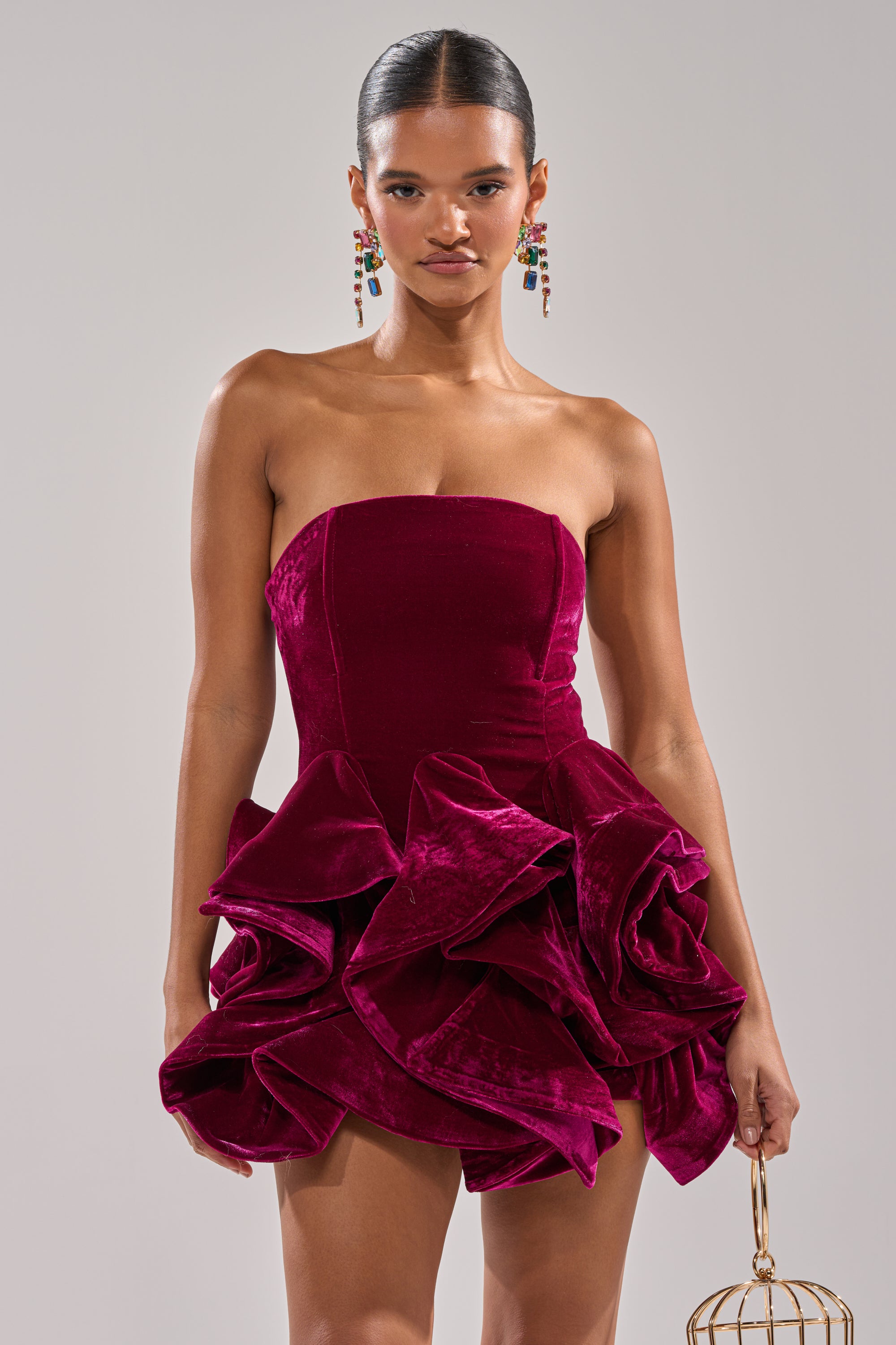 A woman stands against a plain light background wearing the LOVE IS REAL VELVET MINI DRESS, featuring a strapless neckline and dramatic ruffle skirt detailing, paired with statement earrings and a small gold cage handbag.