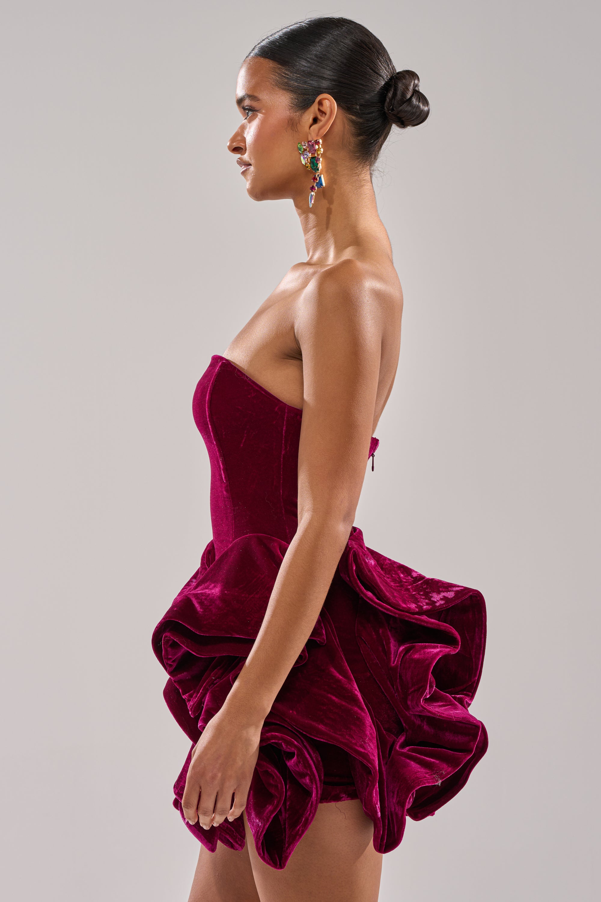 A woman in profile wears the LOVE IS REAL VELVET MINI DRESS, a strapless mini with a sculpted ruffle hem. Her hair is pulled into a sleek bun, and she accessorizes with colorful dangling earrings against a plain light gray backdrop.