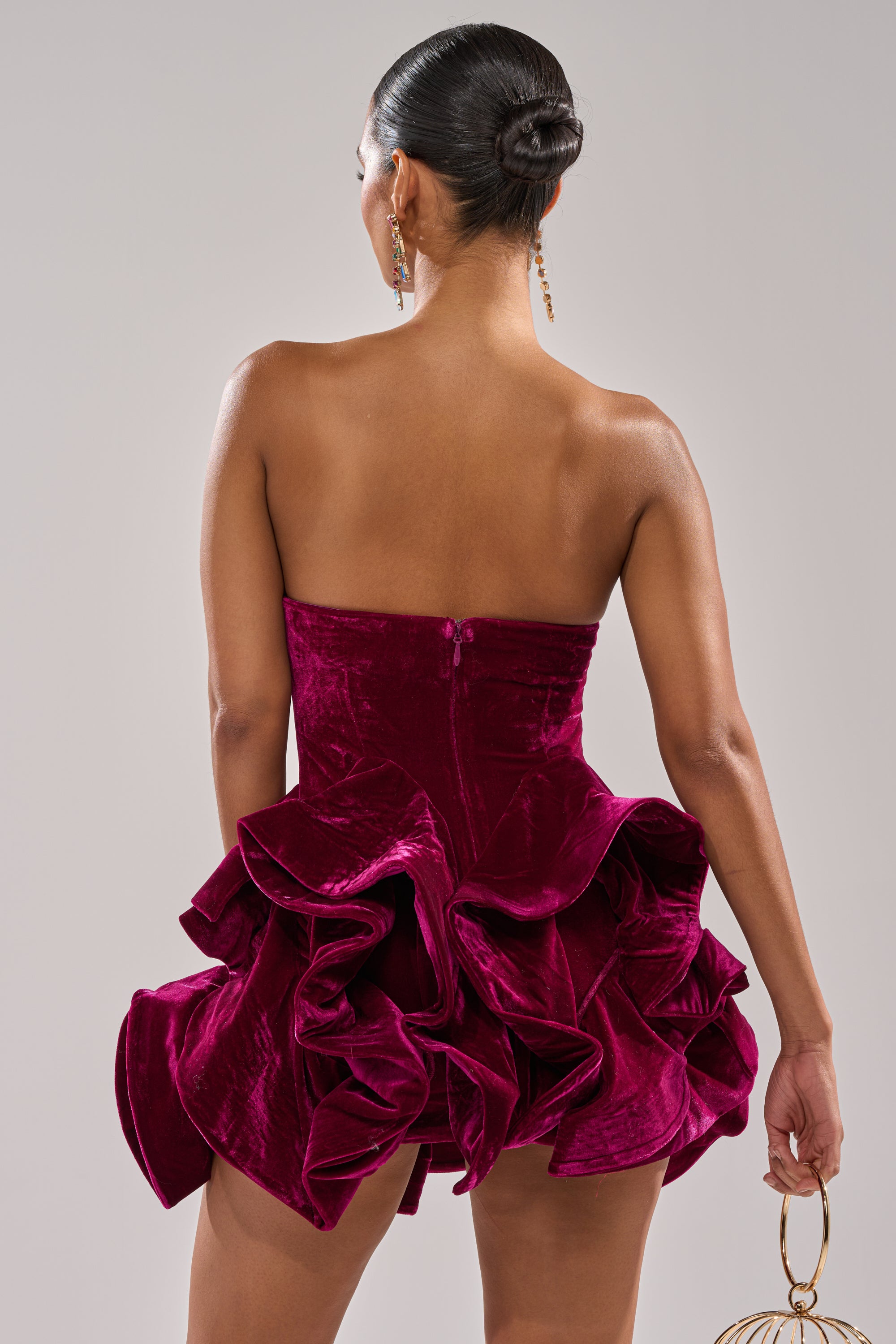 A woman with her hair in a sleek bun is seen from the back wearing the LOVE IS REAL VELVET MINI DRESS, a strapless mini dress with a dramatic ruffle hem. She accessorizes with dangling earrings and a gold circular handbag.