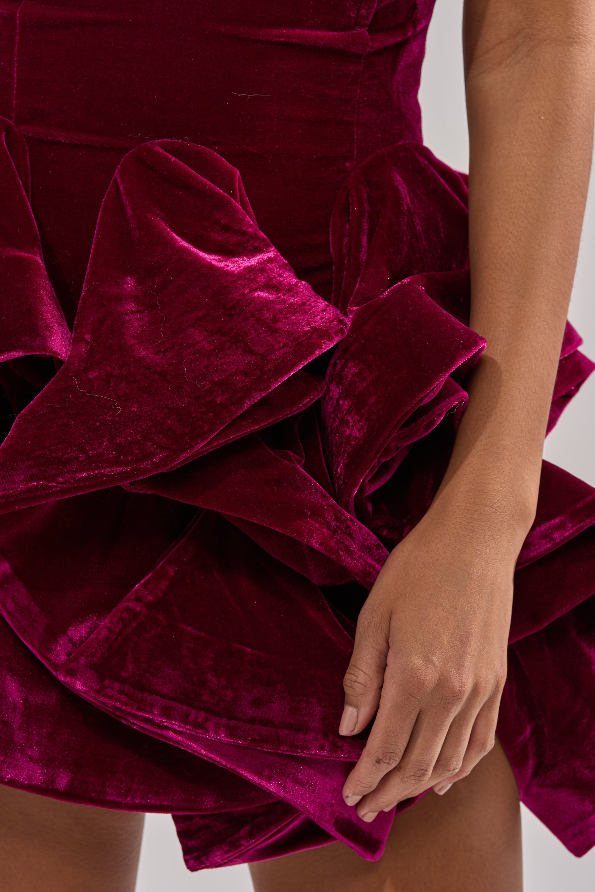 A close-up of a person wearing the LOVE IS REAL VELVET MINI DRESS in deep red, featuring a dramatic layered ruffle hem. Their right arm rests by their side, emphasizing the dress’s rich, glossy velvet texture.