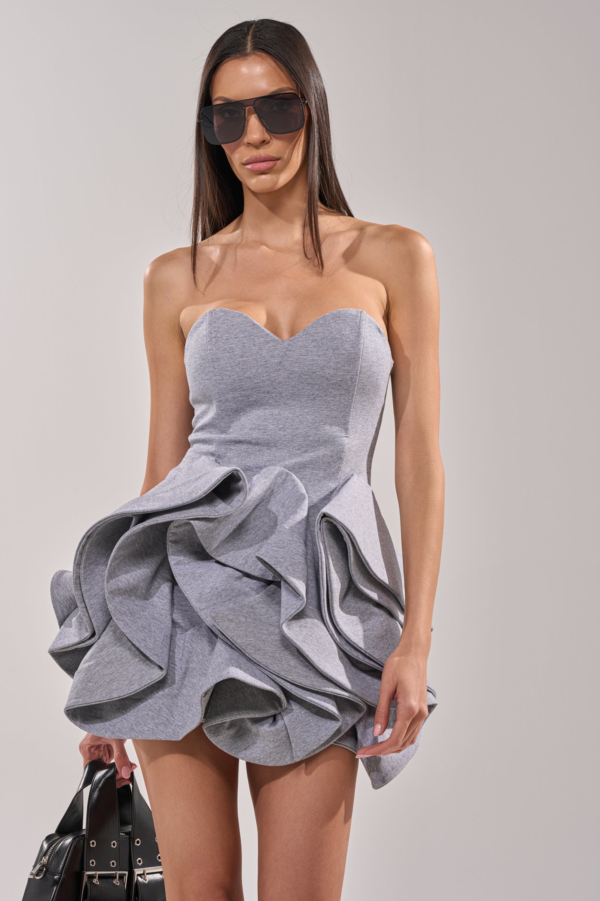 A woman wearing large sunglasses and the CONCRETE BLOOM MINI DRESS—featuring dramatic ruffled layers and a corset-inspired bodice—holds a black handbag while standing against a plain light background. 0