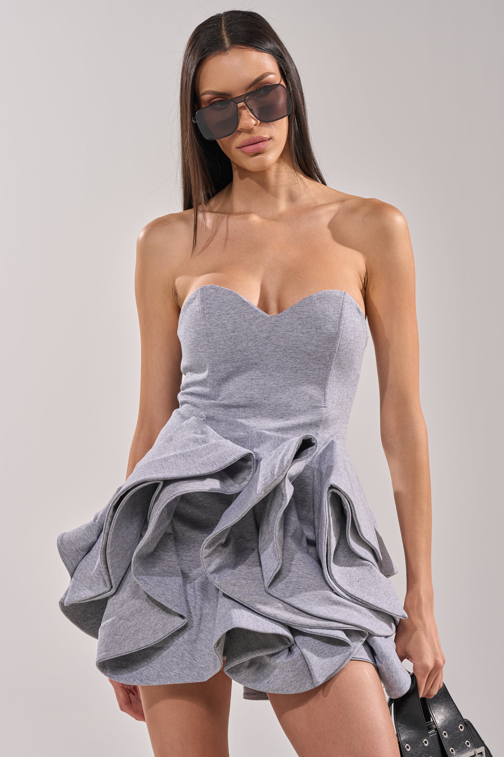 A woman wearing large sunglasses and the CONCRETE BLOOM MINI DRESS, a strapless mini dress with corset-inspired details and ruffled layers, holds a black bag while posing confidently against a plain background. 3
