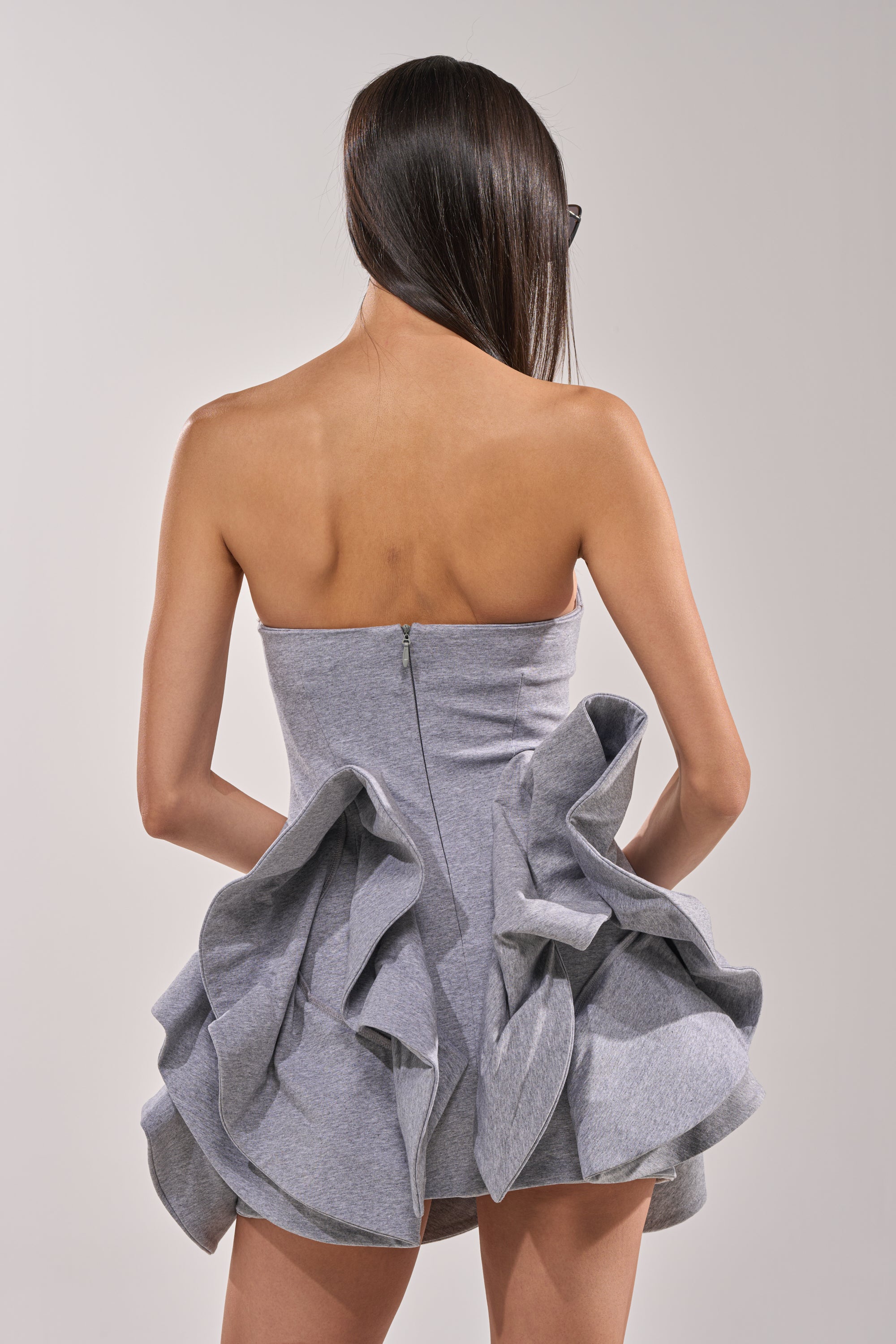 A woman with long, straight hair faces away, wearing the CONCRETE BLOOM MINI DRESS—a strapless mini with a sculptural ruffled bubble hem and corset bodice. The dress exposes her shoulders and upper back, with a zipper running down the back. 5