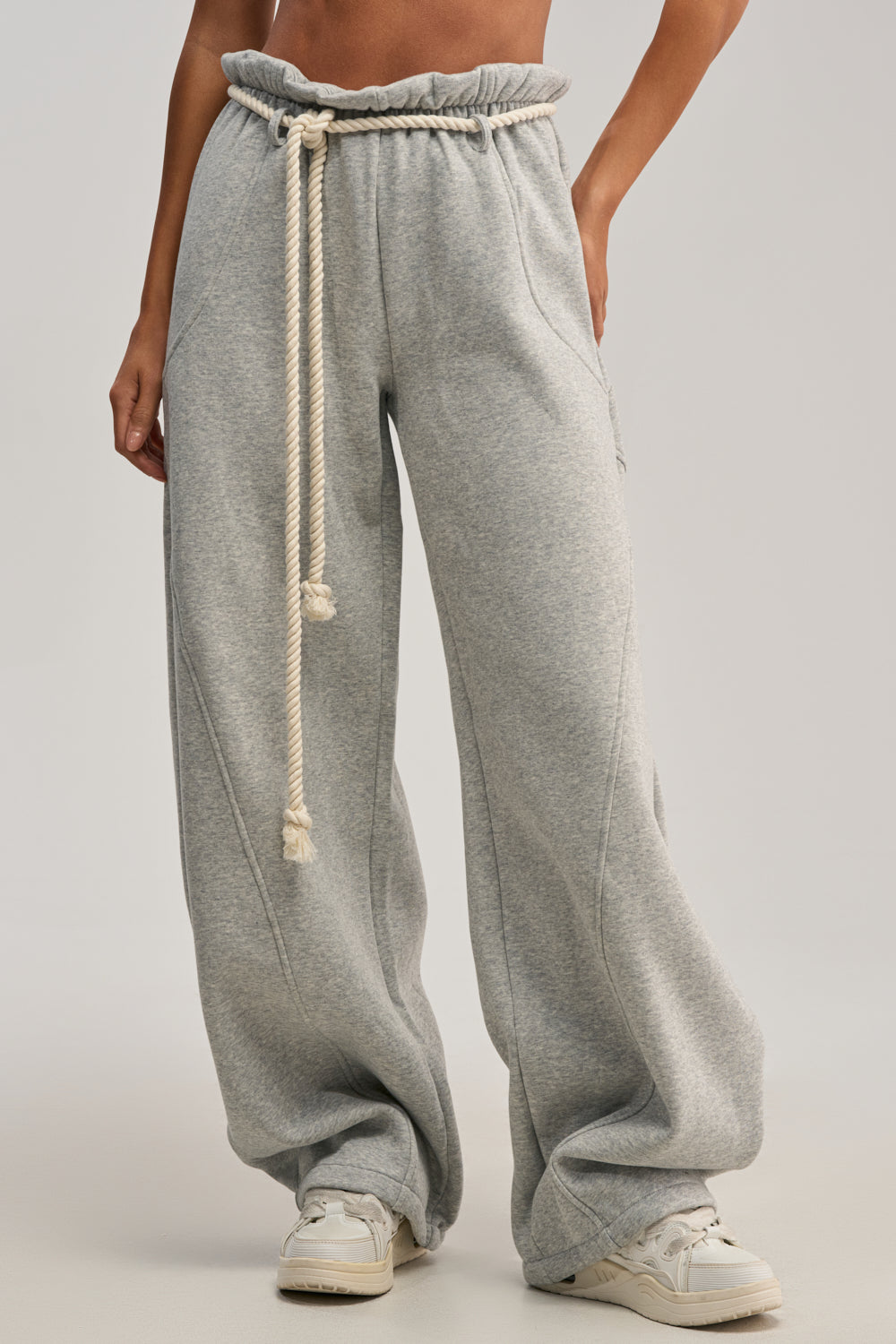 Someone wears IT GIRL WIDE LEG SWEATPANT in light gray, featuring a ruffled waistband and thick rope drawstring, paired with white sneakers. The photo is cropped at the shoulders to showcase the stylish sweatpants.