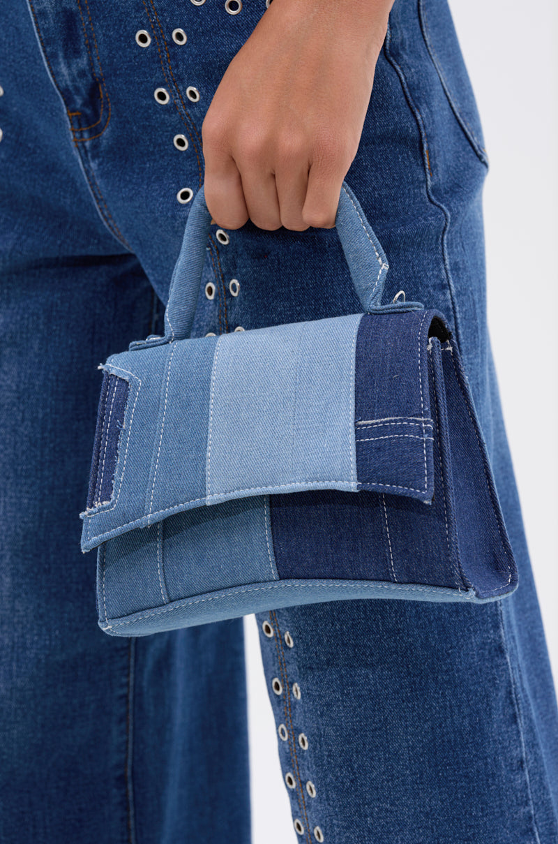 A hand holds the WESTIE PATCHWORK DENIM PURSE, crafted in varied blue denim shades. It comes with a detachable shoulder strap for versatile styling. The person wears blue jeans featuring metal eyelets along the sides. 2