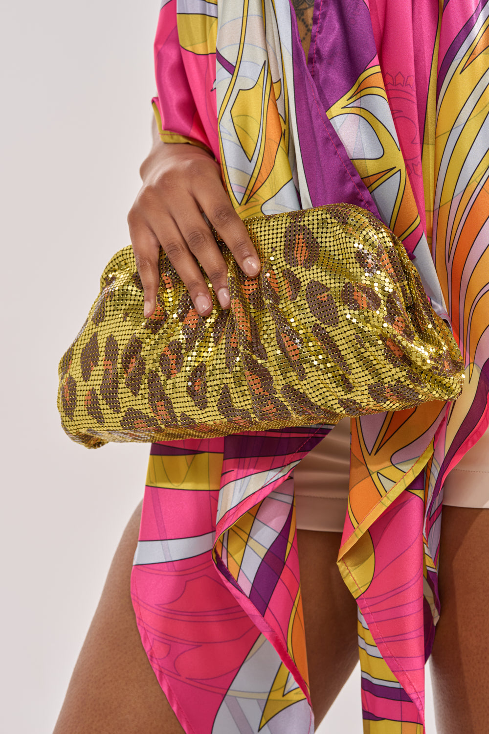 A person in a vibrant, patterned outfit holds the TAKE A TRIP CHAINMAIL CLUTCH, showcasing its gold and brown sequined leopard print against a plain, light-colored background for ultimate Y2K style. 1