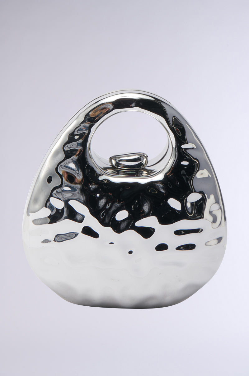 A shiny, metallic abstract sculpture with a rounded triangular shape and central circular opening stands against a plain light gray background. Its reflective, hammered surface evokes the texture of the LOVE MY VIBE HAMMERED CHROME PURSE. 2