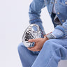 Wearing a light blue denim jacket and jeans, a person sits holding the LOVE MY VIBE HAMMERED CHROME PURSE, showcasing its textured hammered finish and stylish crossbody fit. Only their torso and legs are visible.