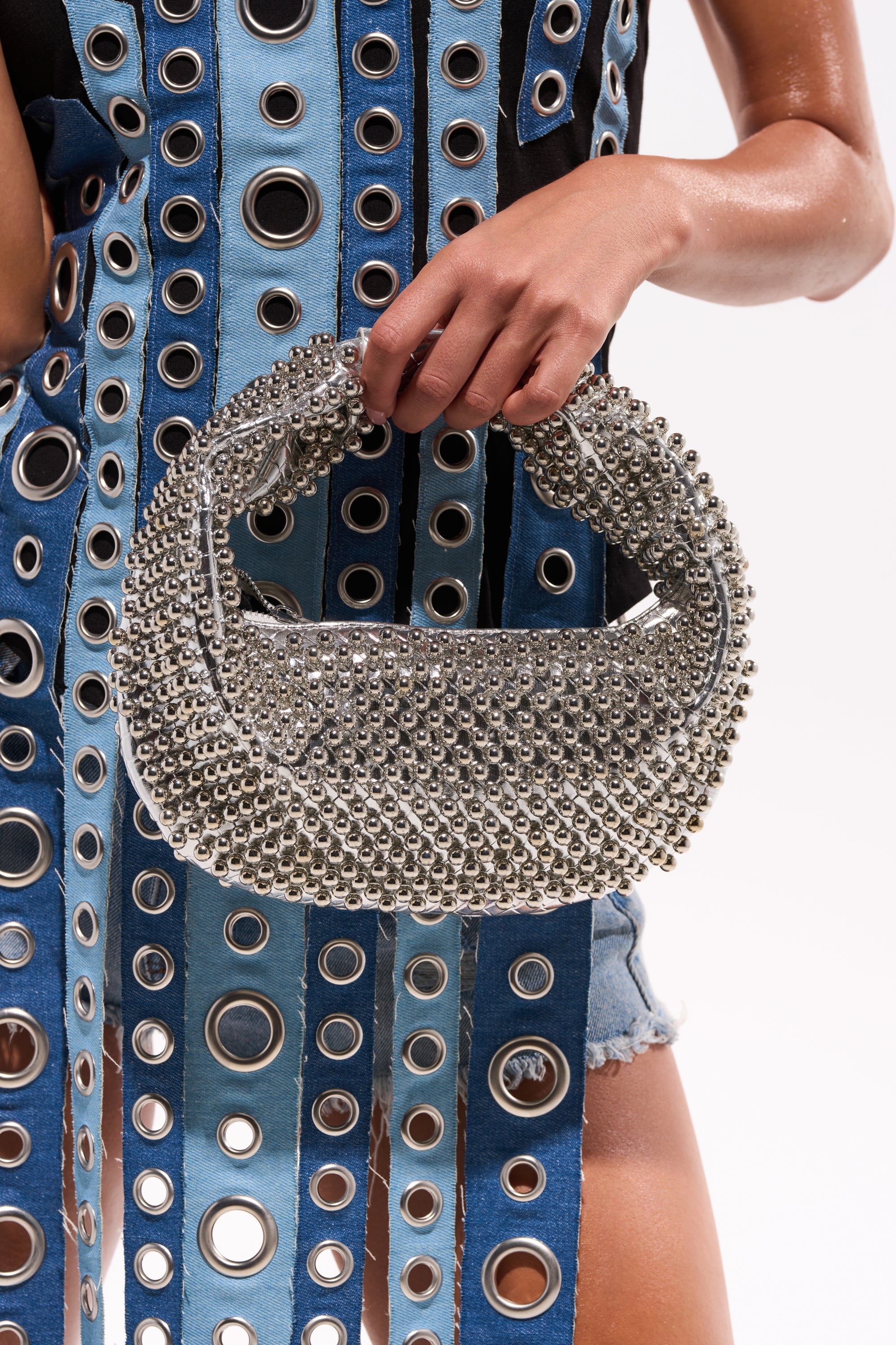 A person in a denim outfit with large metal grommets holds the BEADED BABE PURSE, a crescent-shaped handbag adorned with metallic beads. The white background draws attention to the hand and the distinctive purse. 0