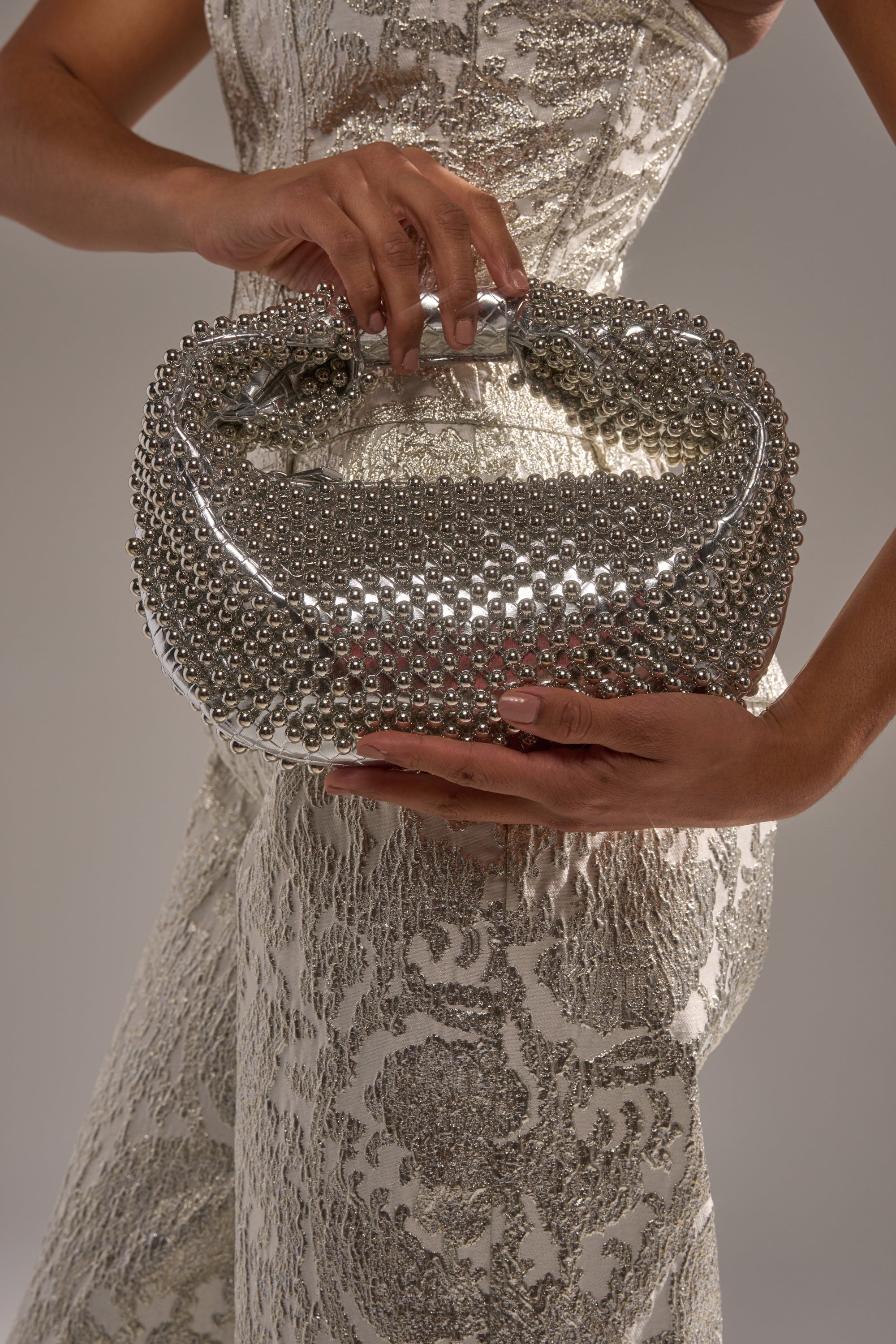 A woman in a textured, metallic-patterned dress holds the BEADED BABE PURSE with both hands, its shimmering silver beads catching the light against a neutral background.