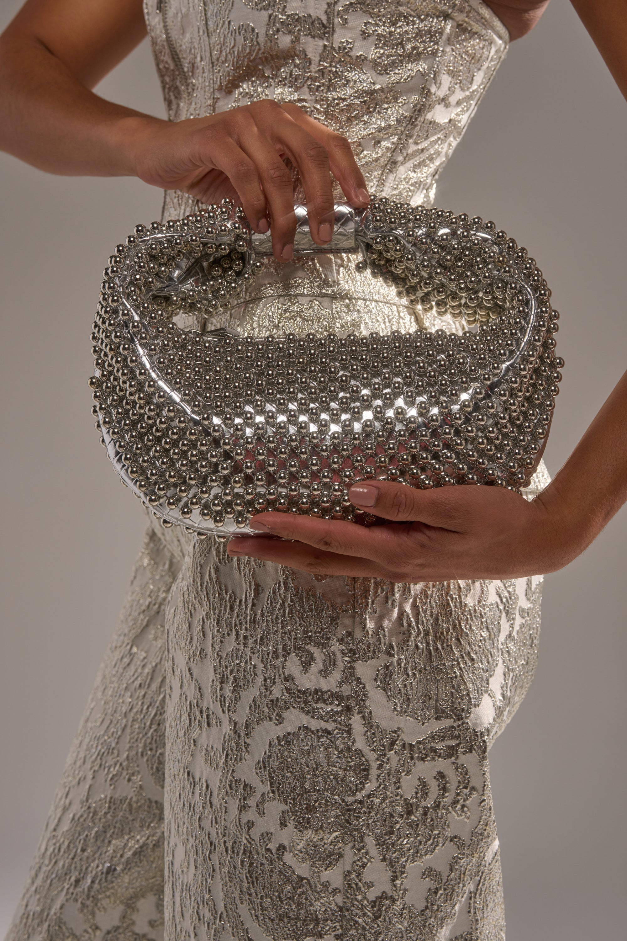 A woman in a textured, metallic-patterned dress holds the BEADED BABE PURSE with both hands, its shimmering silver beads catching the light against a neutral background. 2