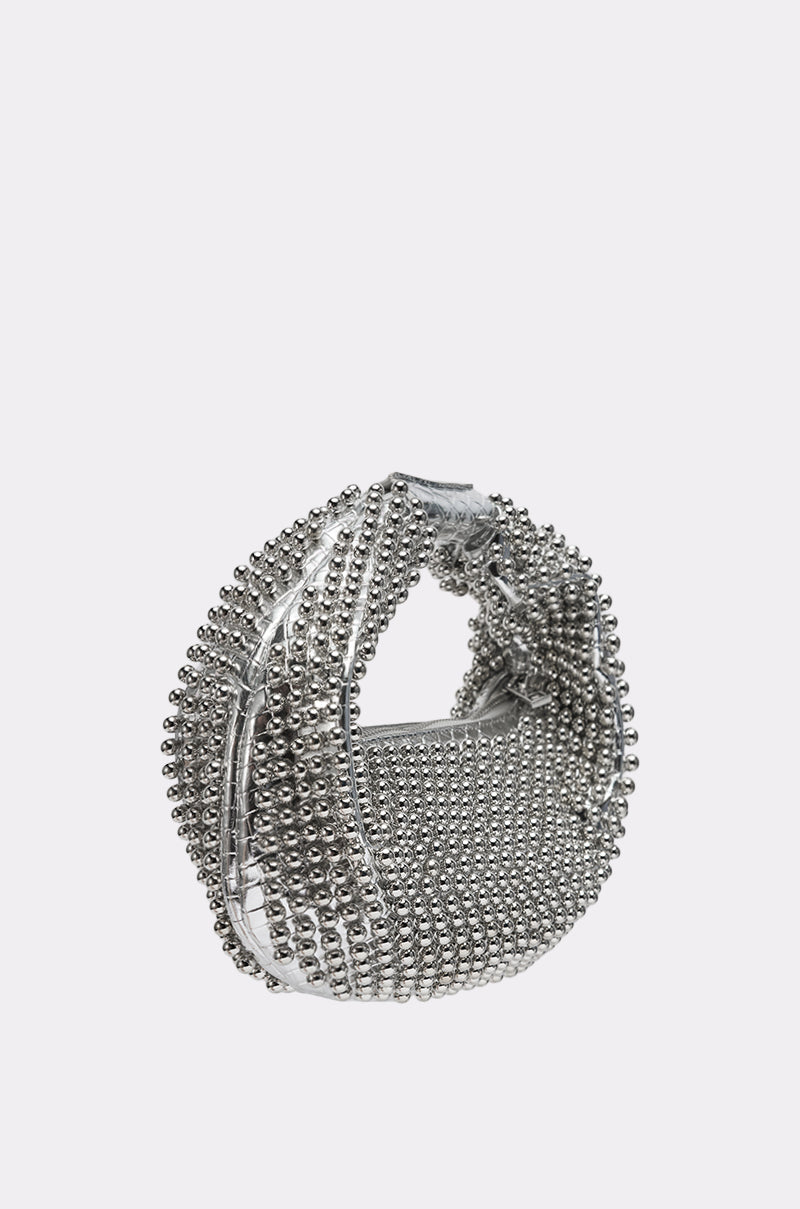 The BEADED BABE PURSE, a small silver crescent-shaped bag with a rounded handle and metallic bead embellishments, is displayed against a plain white background. 3