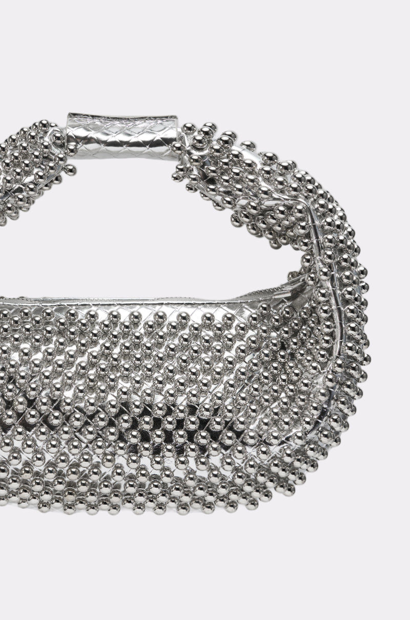 The BEADED BABE PURSE is a silver crescent-shaped bag with shiny rounded studs and a short textured handle, shown against a plain light background. 4