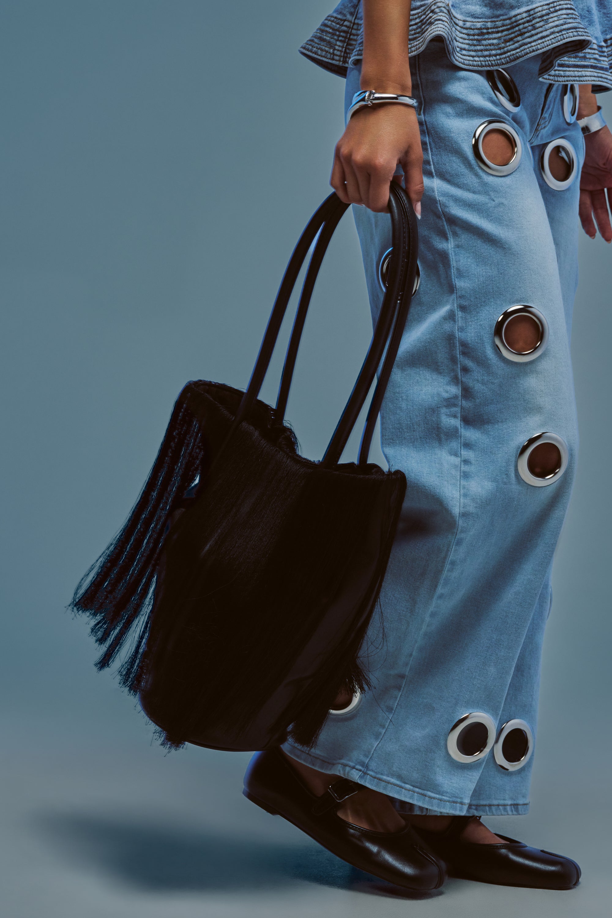 A person, shown from the waist down against a plain background, wears light blue jeans with large grommet holes and black loafers while holding the HAIRY SITUATION TOTE by its handles.