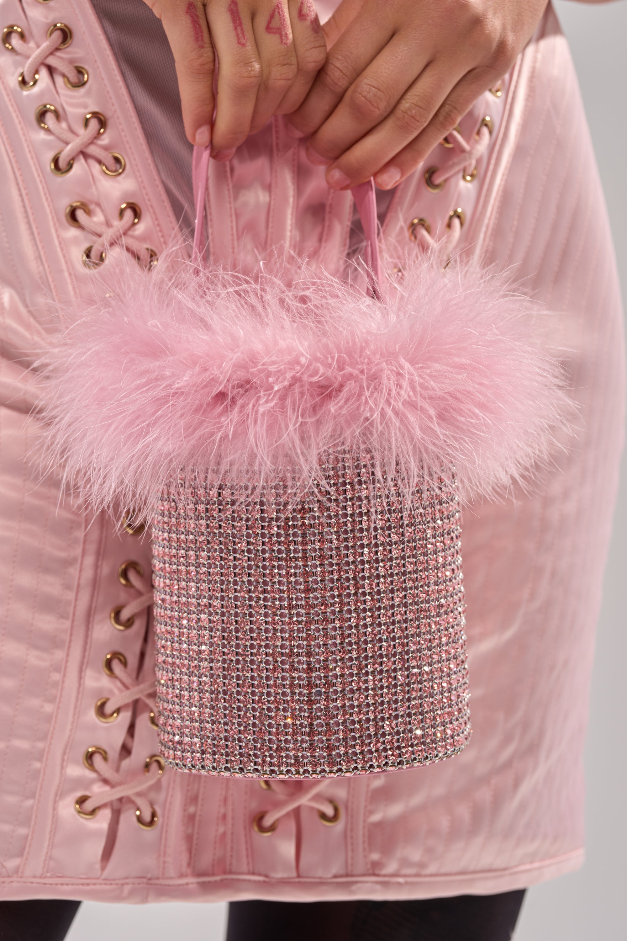 Wearing a light pink dress with lace-up details, a person holds the ELLE PINK FEATHER BLING PURSE—a small rhinestone purse topped with fluffy pink feathers. 4