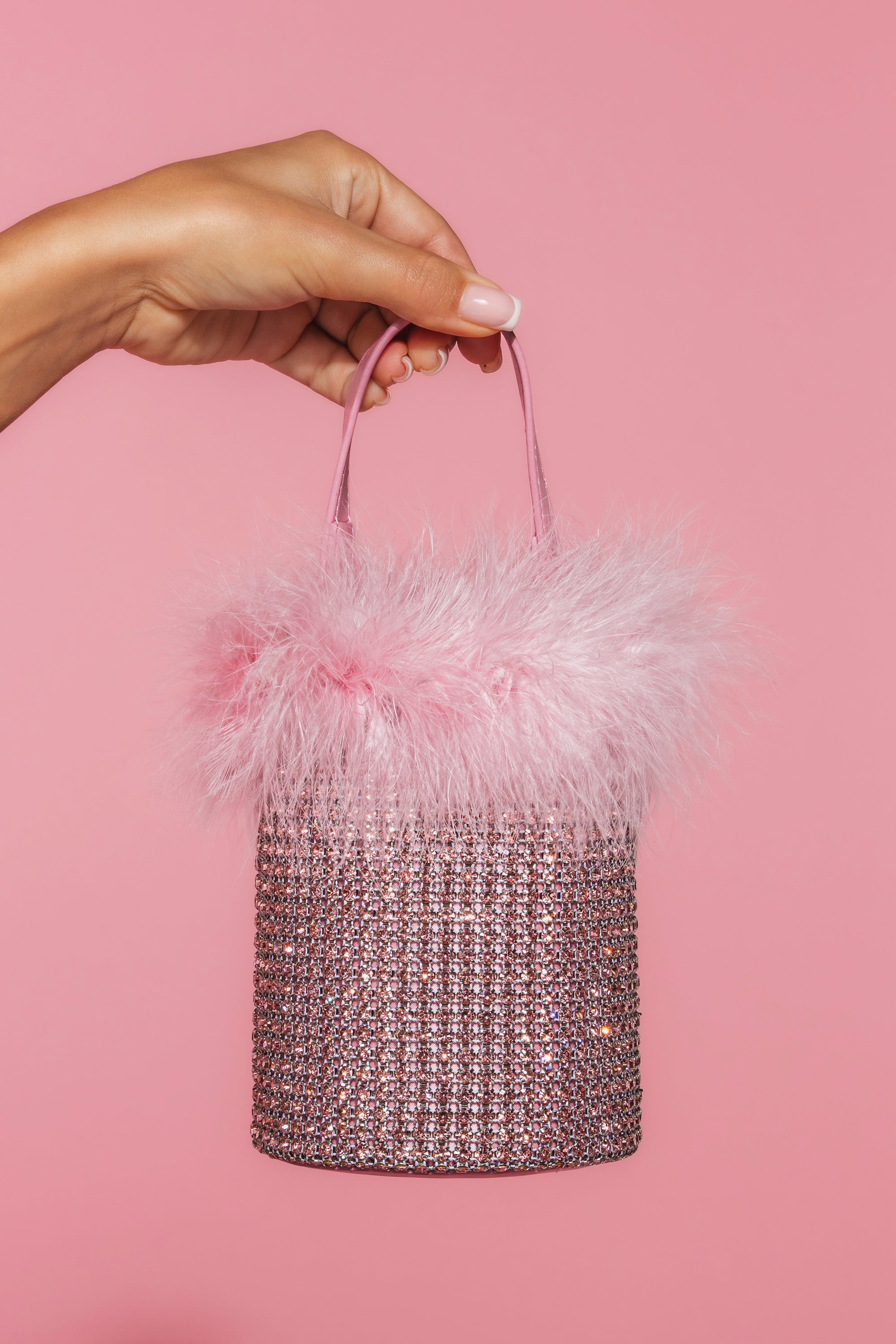 A hand holds the ELLE PINK FEATHER BLING PURSE—pink faux leather, sparkling rhinestones, and fluffy pink feather trim—set against a solid pink background. 2