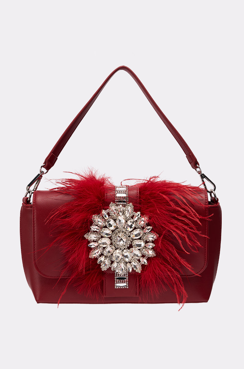 The RED CARPET EMBELLISHED PURSE, crafted from vegan leather, features a top handle, red feathers, and a large ornate silver and crystal brooch at the center. Its bold design stands out against the plain light gray background.