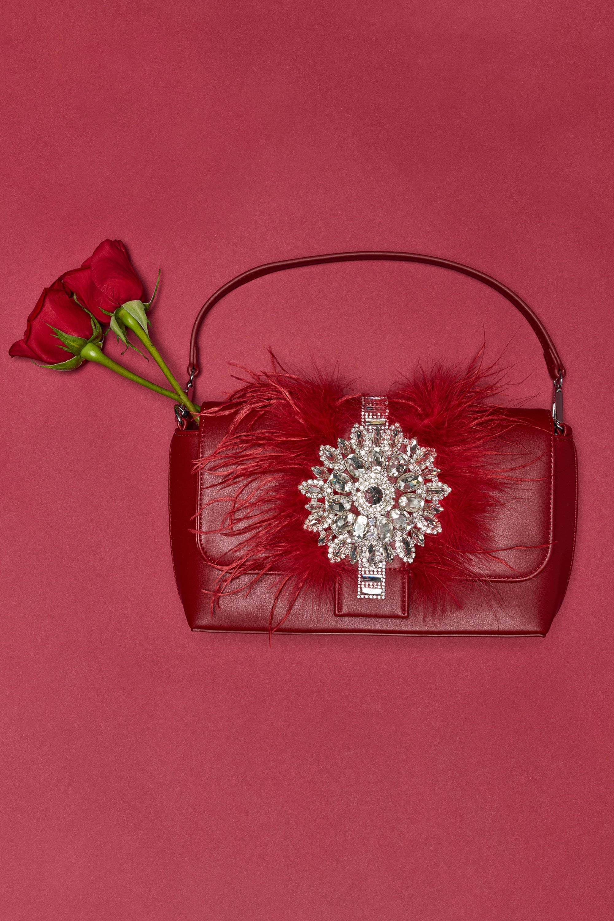 The RED CARPET EMBELLISHED PURSE—vegan leather with a handle, adorned with red feathers and a silver jeweled brooch—rests on a red background. Two red roses are tucked beneath the handle. 0
