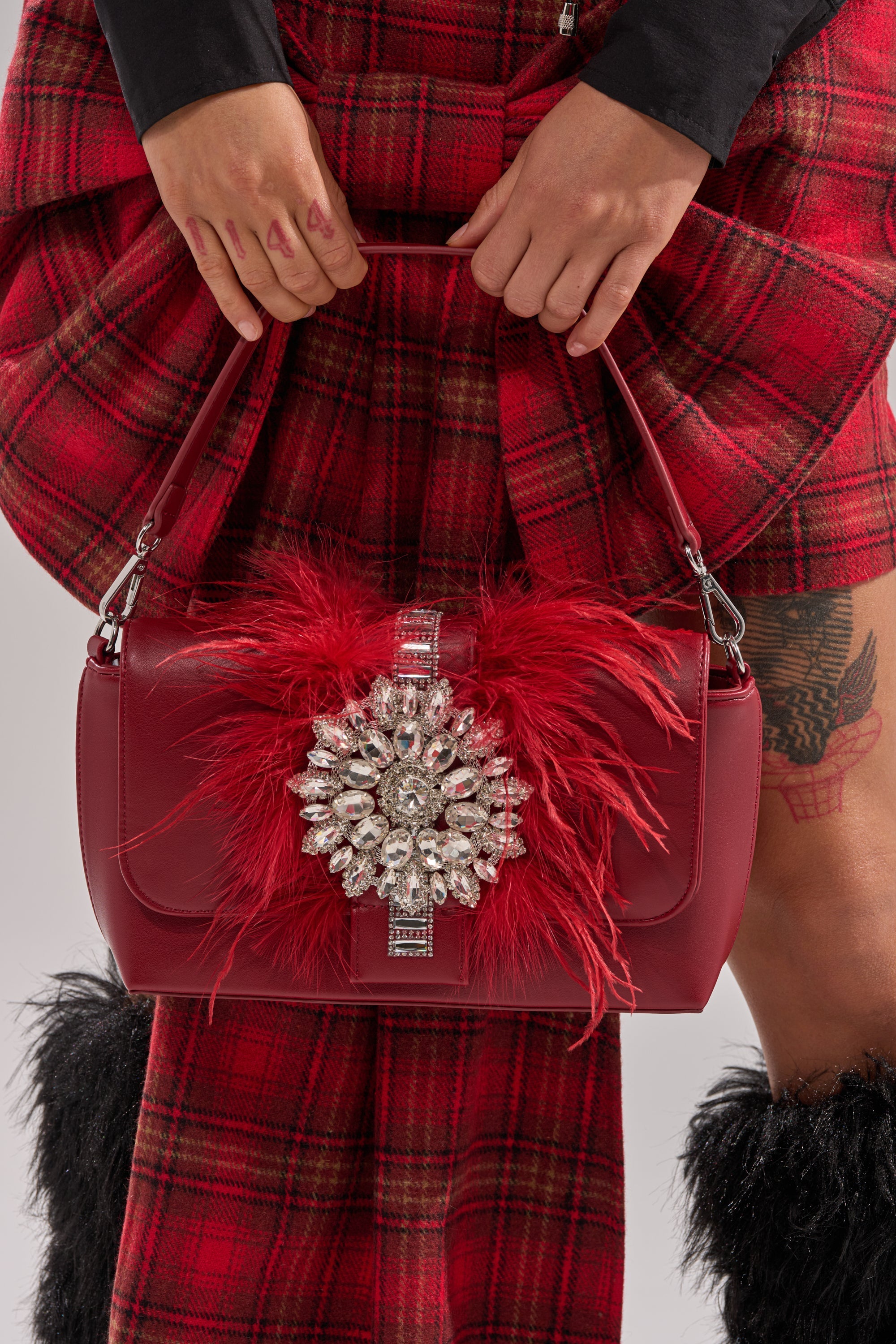 A person in red plaid holds the RED CARPET EMBELLISHED PURSE featuring red feathers and a jeweled accent. Paired with a leg tattoo and furry black boots, this look is perfect for showcasing a statement accessory. 2