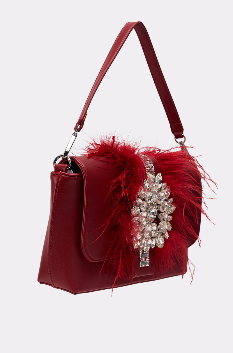 The RED CARPET EMBELLISHED PURSE features a top handle, vegan leather exterior, red feather details, and a striking silver rhinestone brooch on the front flap.