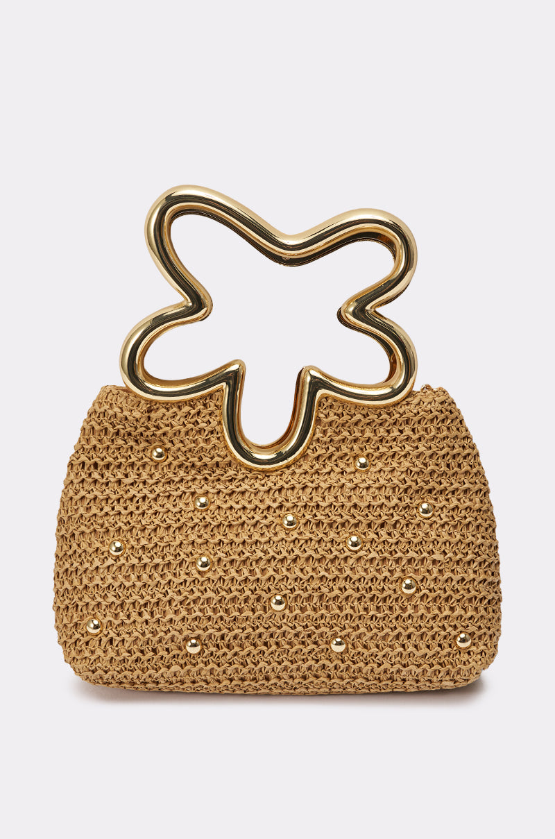 The FLOWER POWER PURSE is a tan woven straw handbag with gold studs and a unique gold abstract flower handle. It features a gold chain crossbody strap, making it the ideal summer accessory against any plain light background.