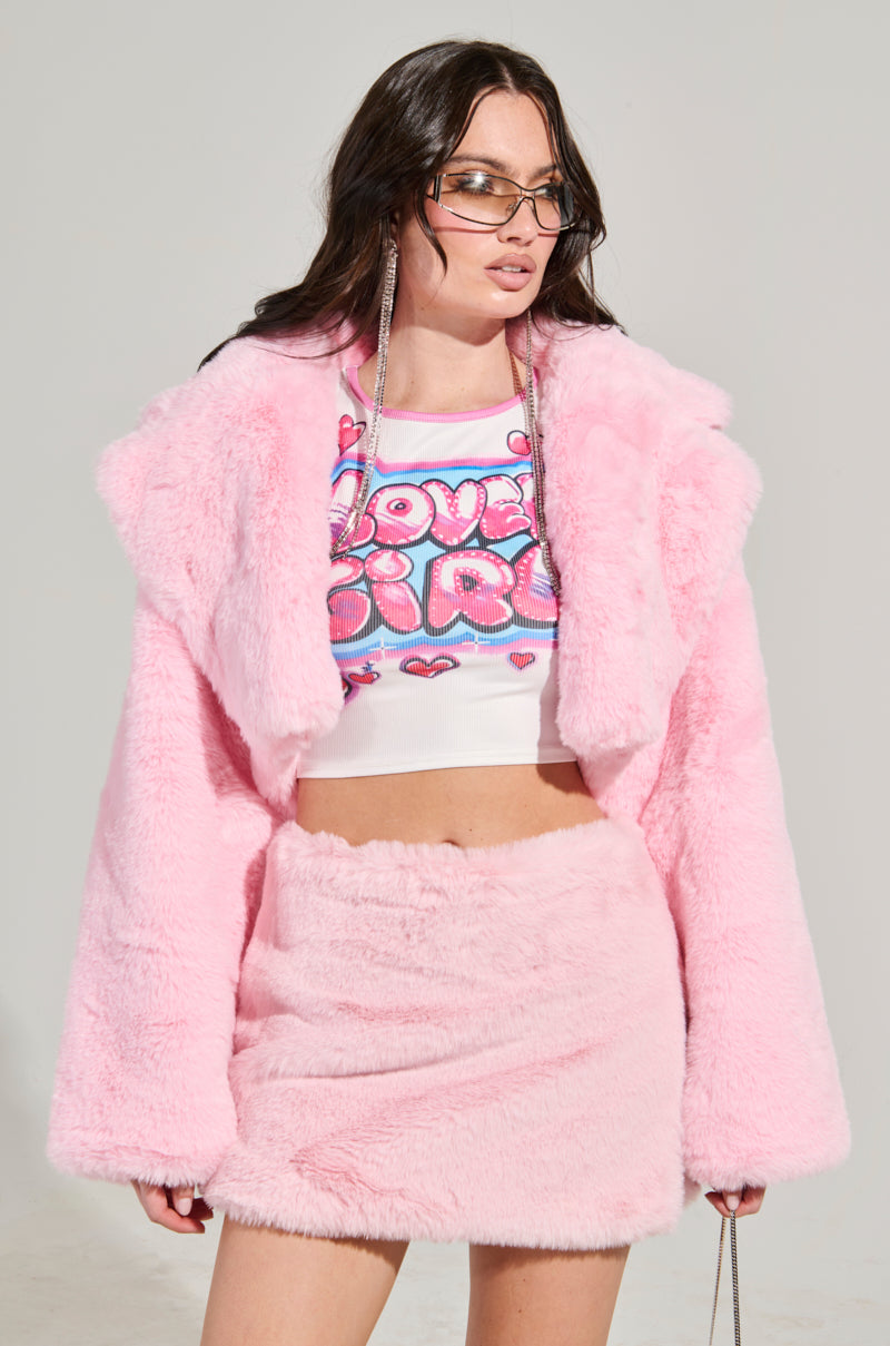 A woman confidently poses in a POPPY CROPPED FUR COAT IN LIGHT PINK, paired with a skirt, a "Love Girl" graphic top, and sunglasses.