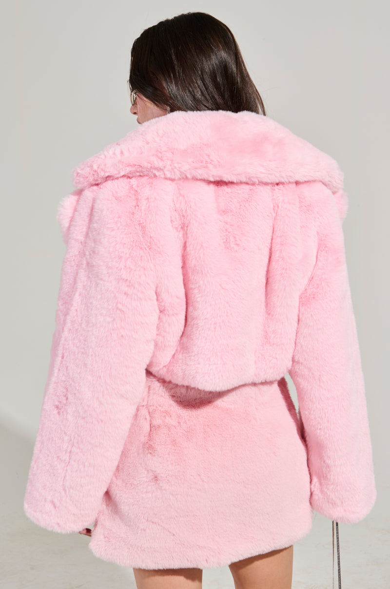 A person with long dark hair is facing away, showcasing the plush POPPY Cropped Fur Coat in Light Pink, featuring a wide collar and belt. The background is plain and light-colored.