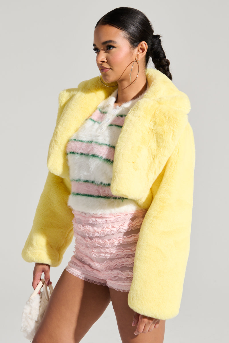Wearing the POPPY CROPPED FUR COAT IN YELLOW with an oversized lapel collar, the person stands out as they layer it over a striped fuzzy top and pair it with pink lace shorts. They complete their bold look with large hoop earrings and have their hair elegantly styled in a sleek ponytail against a plain light gray background.