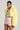 Wearing the POPPY CROPPED FUR COAT IN YELLOW, a person poses confidently in an oversized lapel collar fur coat over a striped fuzzy top and pink ruffled shorts. Large hoop earrings dangle as their hair is pulled back, standing out against the plain, light-colored background.