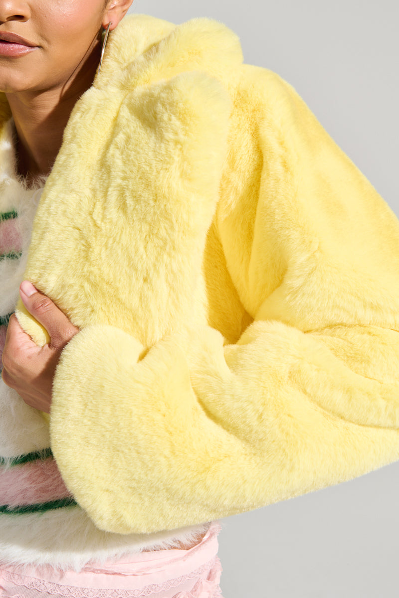 Close-up of an individual wearing the vibrant POPPY CROPPED FUR COAT IN YELLOW, featuring an oversized lapel collar. The shot captures the texture of the coat, with hands partially visible as they hold it closed against a neutral gray backdrop, highlighting its striking color and cozy charm.
