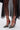 A person in a sheer black skirt wears AZALEA WANG LYSANDER BLACK MULTI PUMP STILETTOS with patent faux leather, chunky gemstone embellishments, and red ankle straps, posed against a plain white background.
