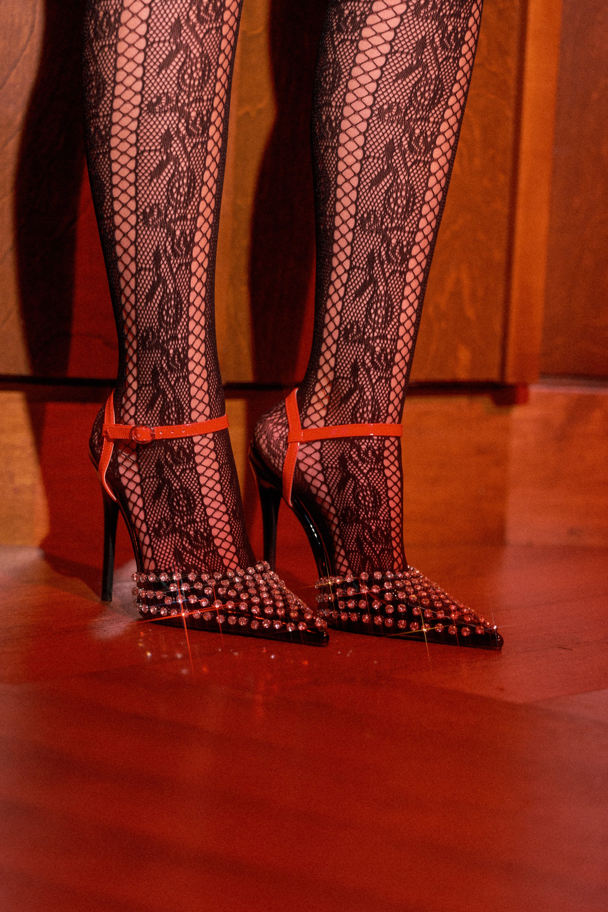 A person stands on a wooden floor wearing black lace tights and striking AZALEA WANG LYSANDER BLACK MULTI PUMP STILETTO heels, adorned with silver studs and red ankle straps, illuminated by warm lighting.