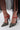 A close-up of someone wearing the AZALEA WANG LYSANDER BLACK MULTI PUMP STILETTO with patent faux leather, chunky gemstone embellishments, sheer black stockings, and a flowy black skirt.