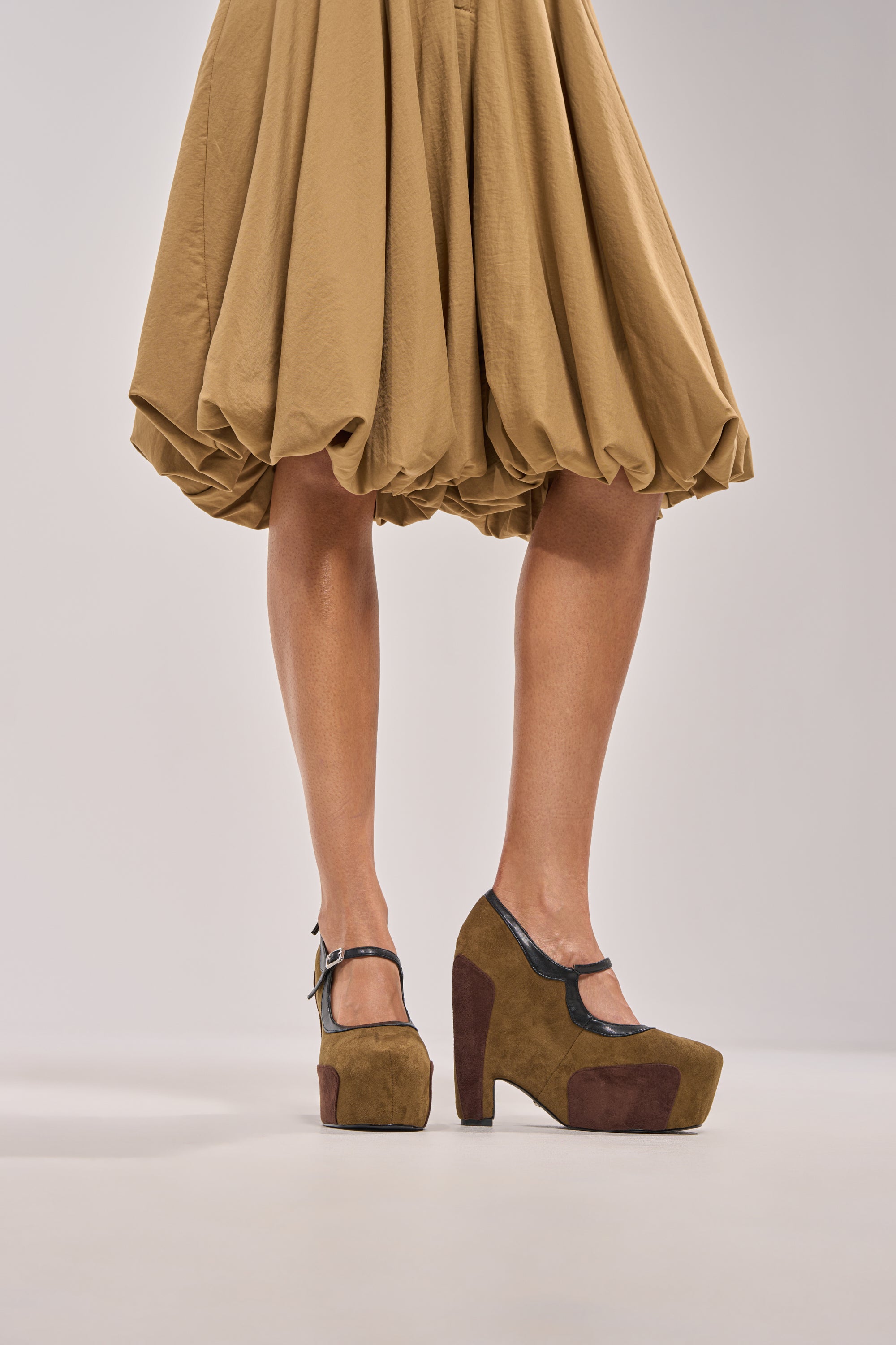 A person stands against a plain background, spotlight on their legs and shoes: the AZALEA WANG LYTHA OLIVEMARY JANE PUMP, featuring brown platform soles and black straps with a subtle Mary Jane-inspired design. 1