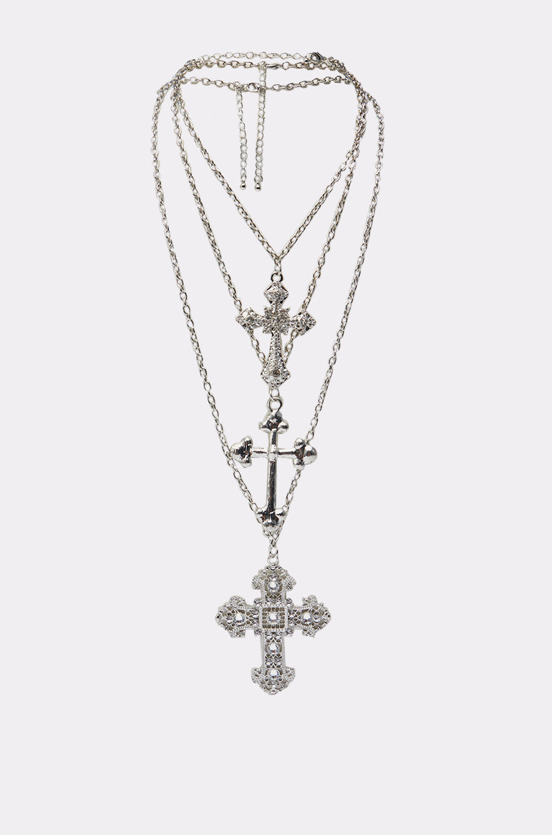 The CROSS MY HEART NECKLACE showcases three silver layered chains with unique cross pendants—one accented with rhinestones—elegantly displayed on a plain white background.