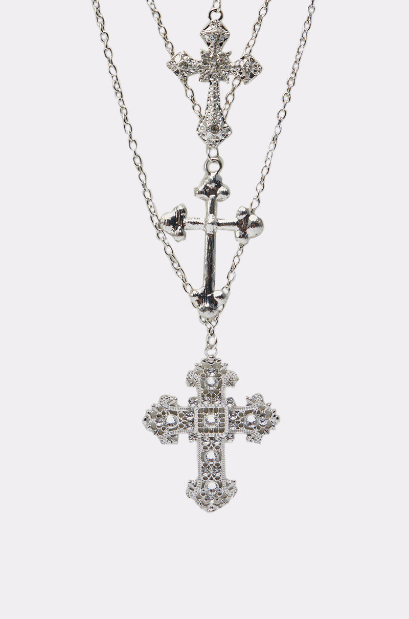 The CROSS MY HEART NECKLACE features three layered chains, each with an ornate cross pendant showcasing intricate designs, displayed on a plain white background.